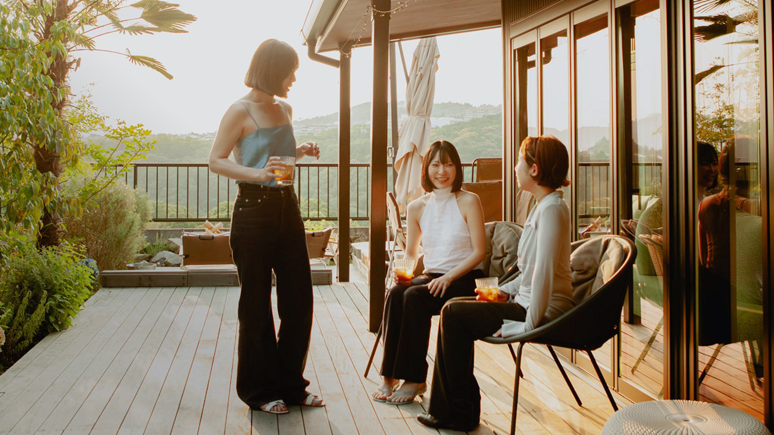 3人の女性が、夕暮れの自然豊かなバルコニーで飲み物を片手に談笑する様子