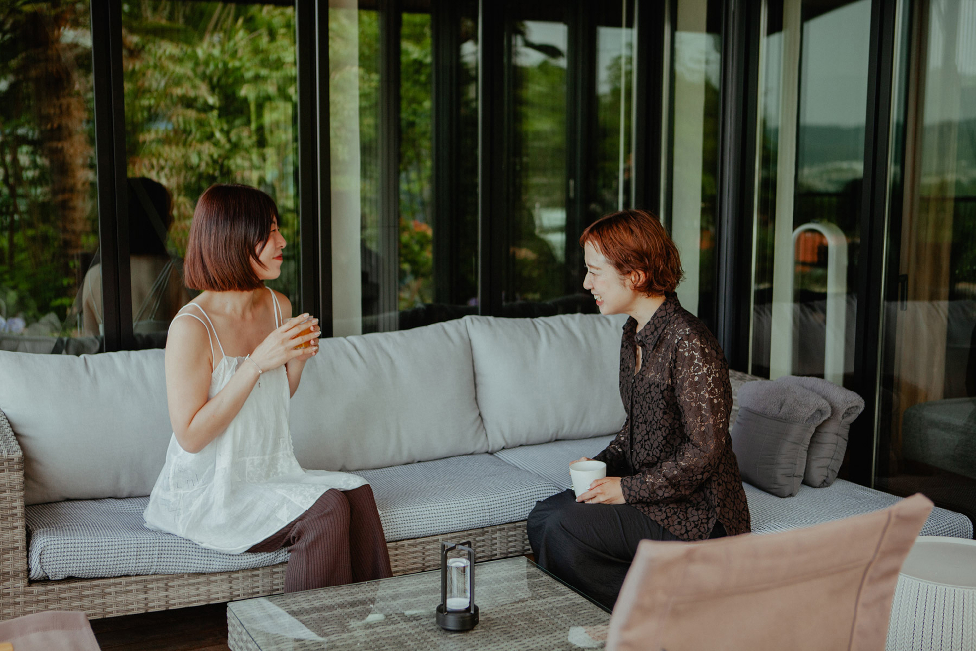 二人の女性が早朝のテラスで、ソファに腰掛けてコーヒー片手に談笑している様子