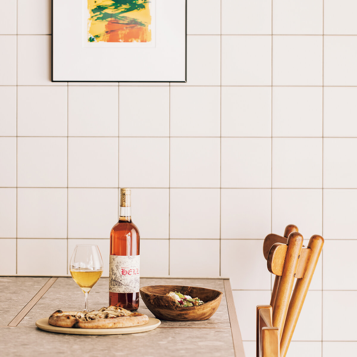 Wine bottle with a glass of white wine, pizza on a board, and a wooden salad bowl on a kitchen counter against white tiled wall with a wooden chair nearby.