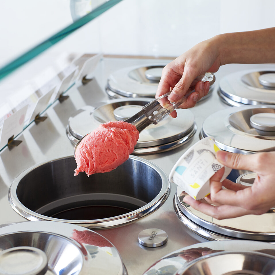 Hand scooping bright pink sorbet from a stainless steel ice-cream display with a metal scoop in a shop setting.