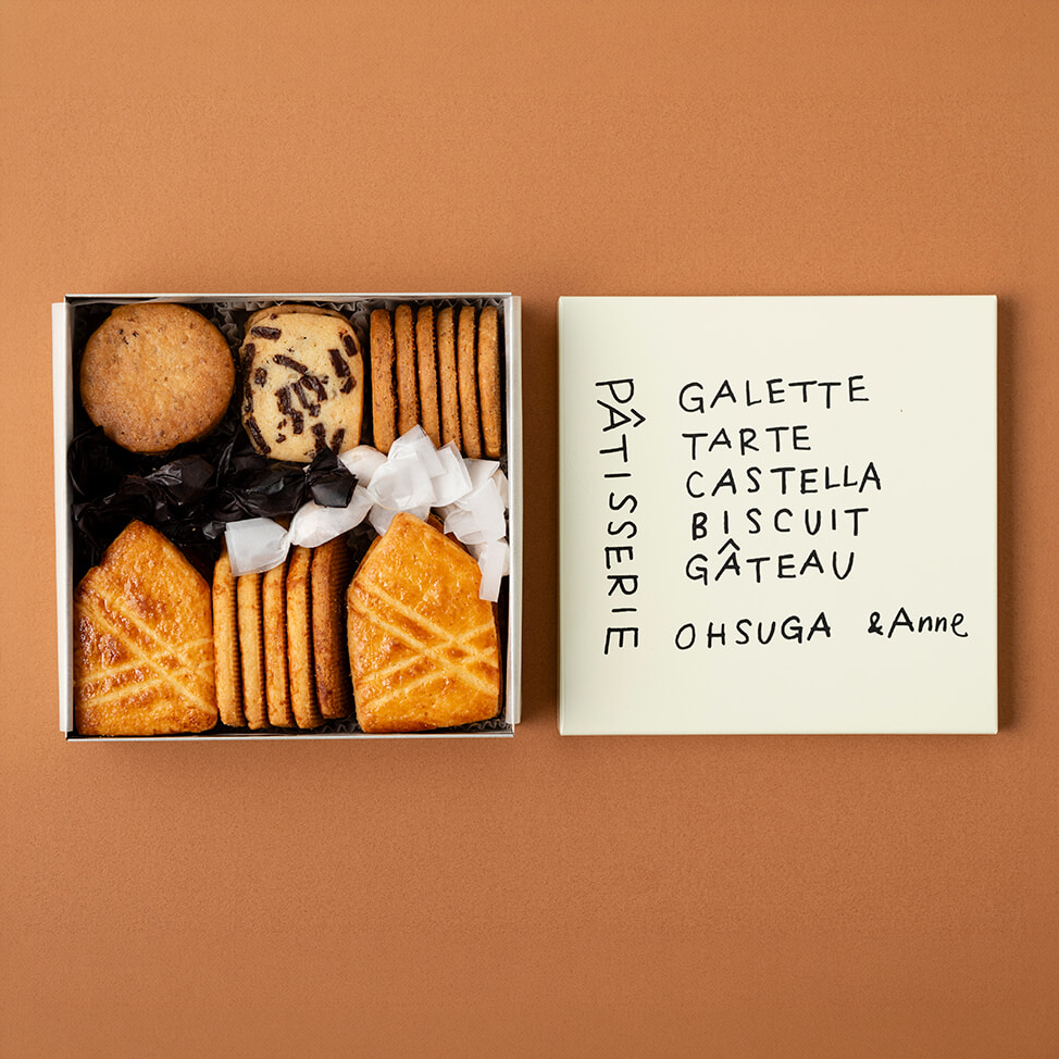Open box of assorted cookies with round, chocolate-chip, and rectangular biscuits, beside a cream box with handwritten 'PATISSERIE' text on a peachy background.