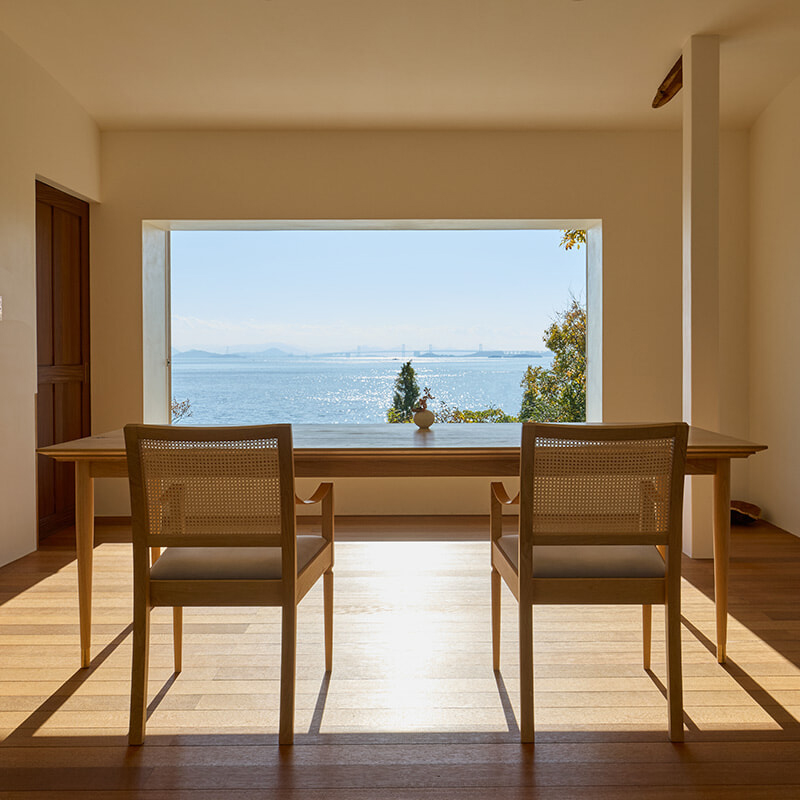 Wooden table with two chairs facing a large window overlooking the sea and distant bridge, in a bright minimalist room.