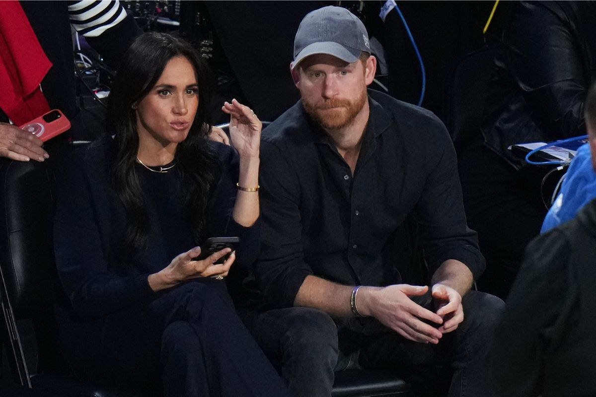 Meghan Markle and Prince Harry sit together; Meghan gestures with a phone in hand, Harry wearing a gray cap.