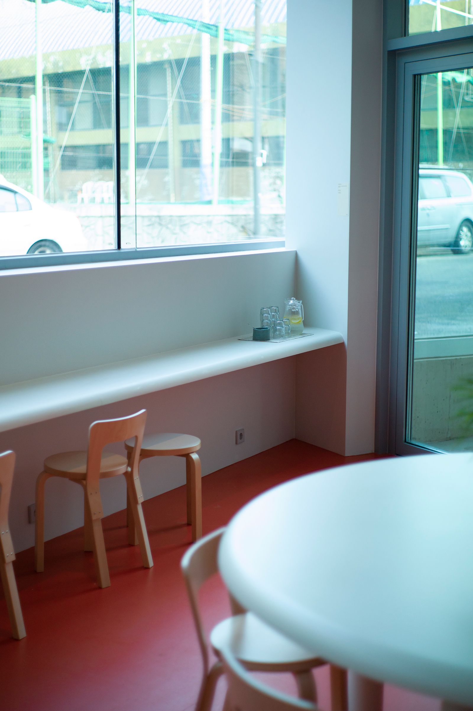 Bright interior with a white windowsill along a glass wall, light wood stools, and a round white table on a red floor; a glass jug on the sill.