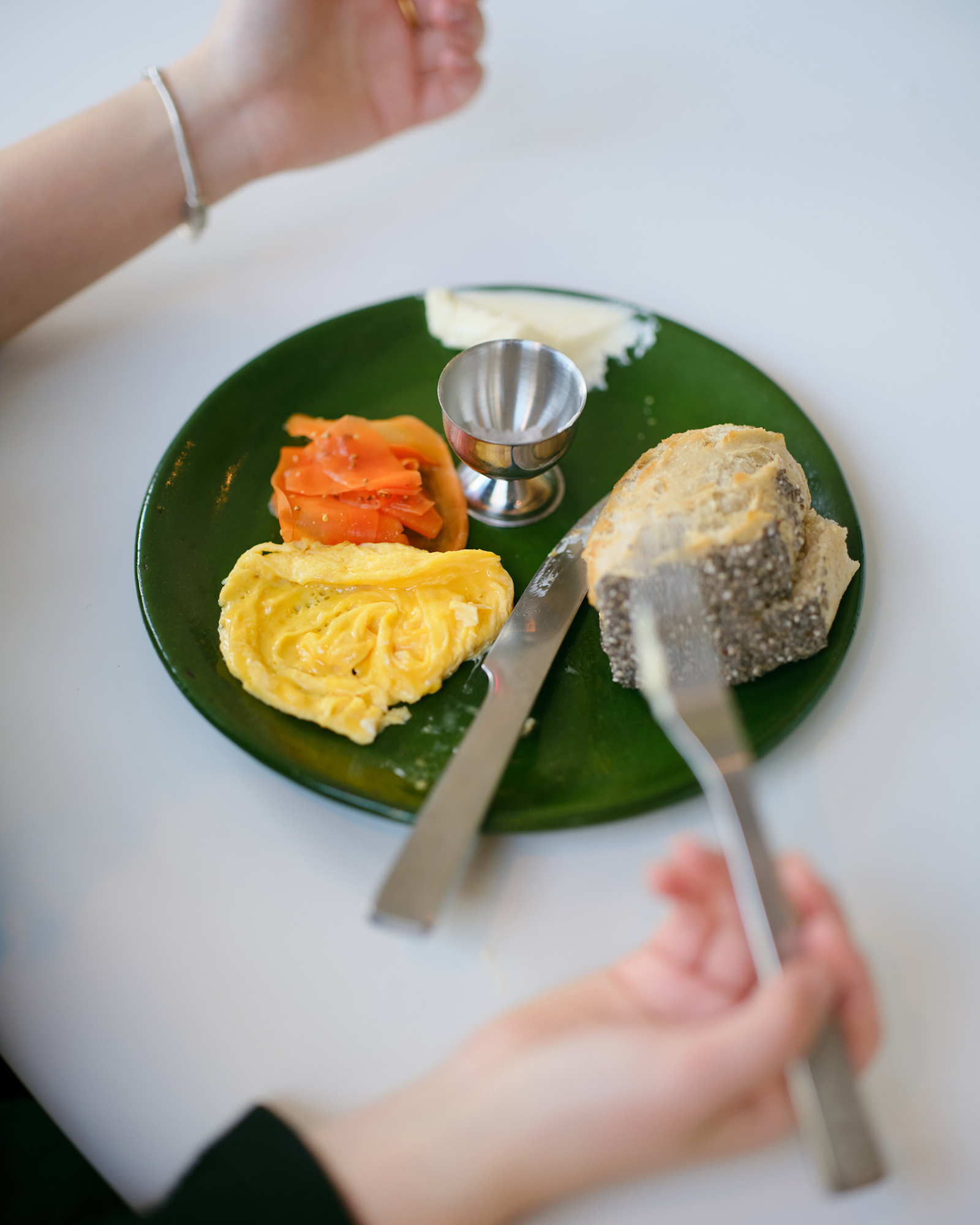 Person's hands cutting into bread on a green plate with scrambled eggs, smoked salmon, a butter pat, and a small metal egg cup; breakfast scene.