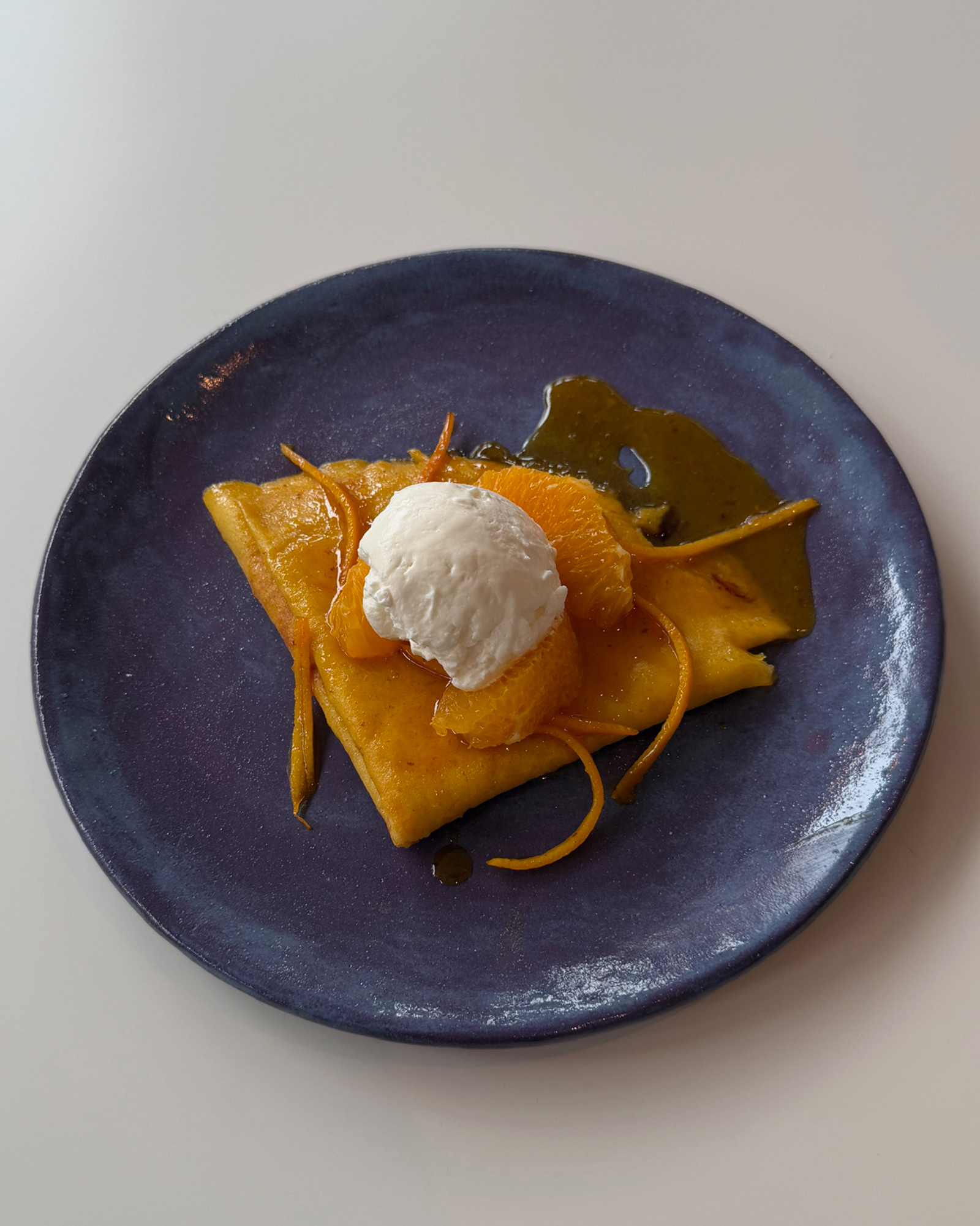Dessert on a blue plate: a folded crepe with orange segments, a scoop of vanilla ice cream, and caramel drizzle.