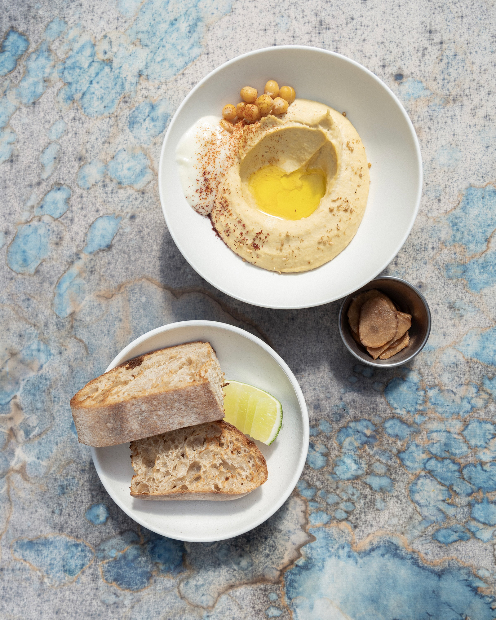 Top-down view of hummus with olive oil, chickpeas on a plate, served with crusty bread slices and a lime wedge; small cup of nut butter beside them.