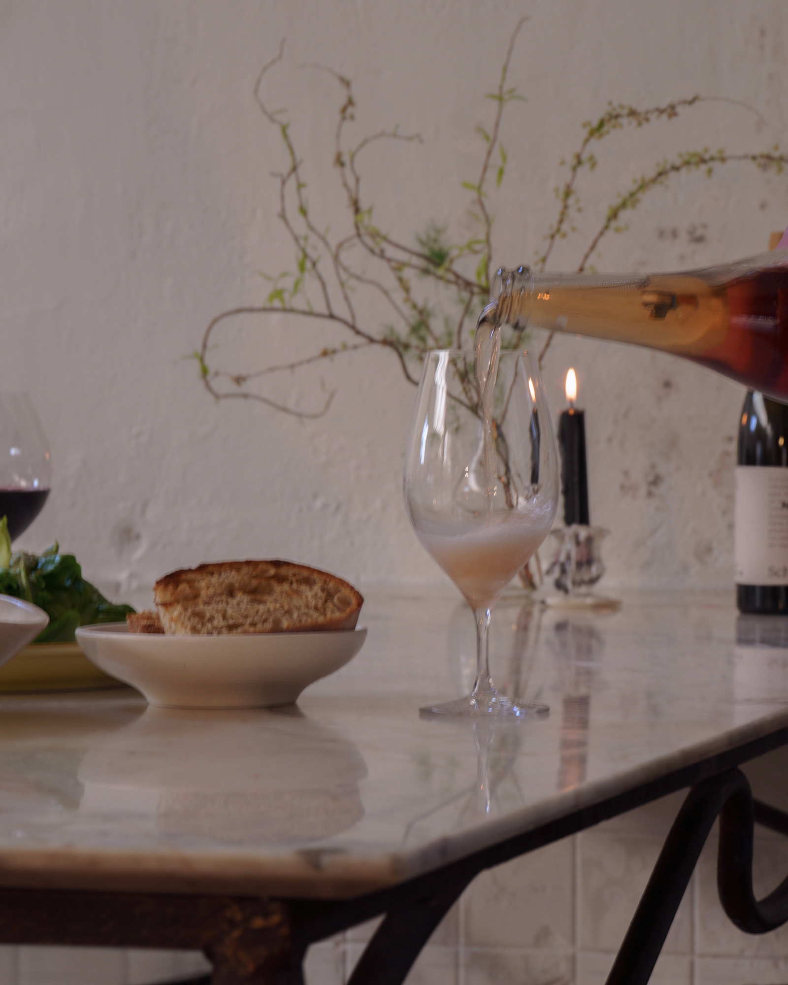 Wine being poured into a stemmed glass on a marble table, with bread, a candle, and a wine bottle in the background.