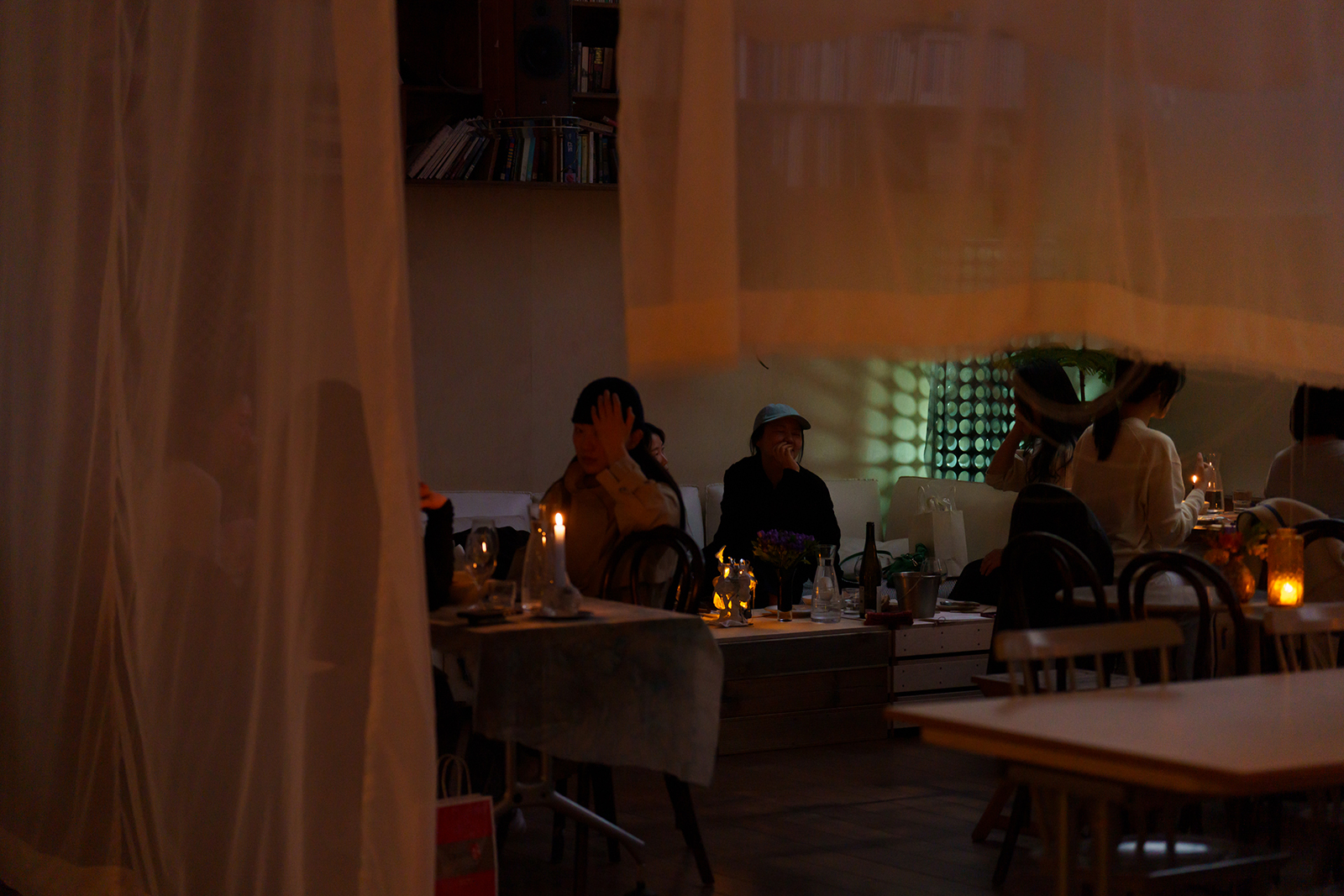 Dimly lit dining room with candles on tables and guests seated, sheer curtains framing the scene.