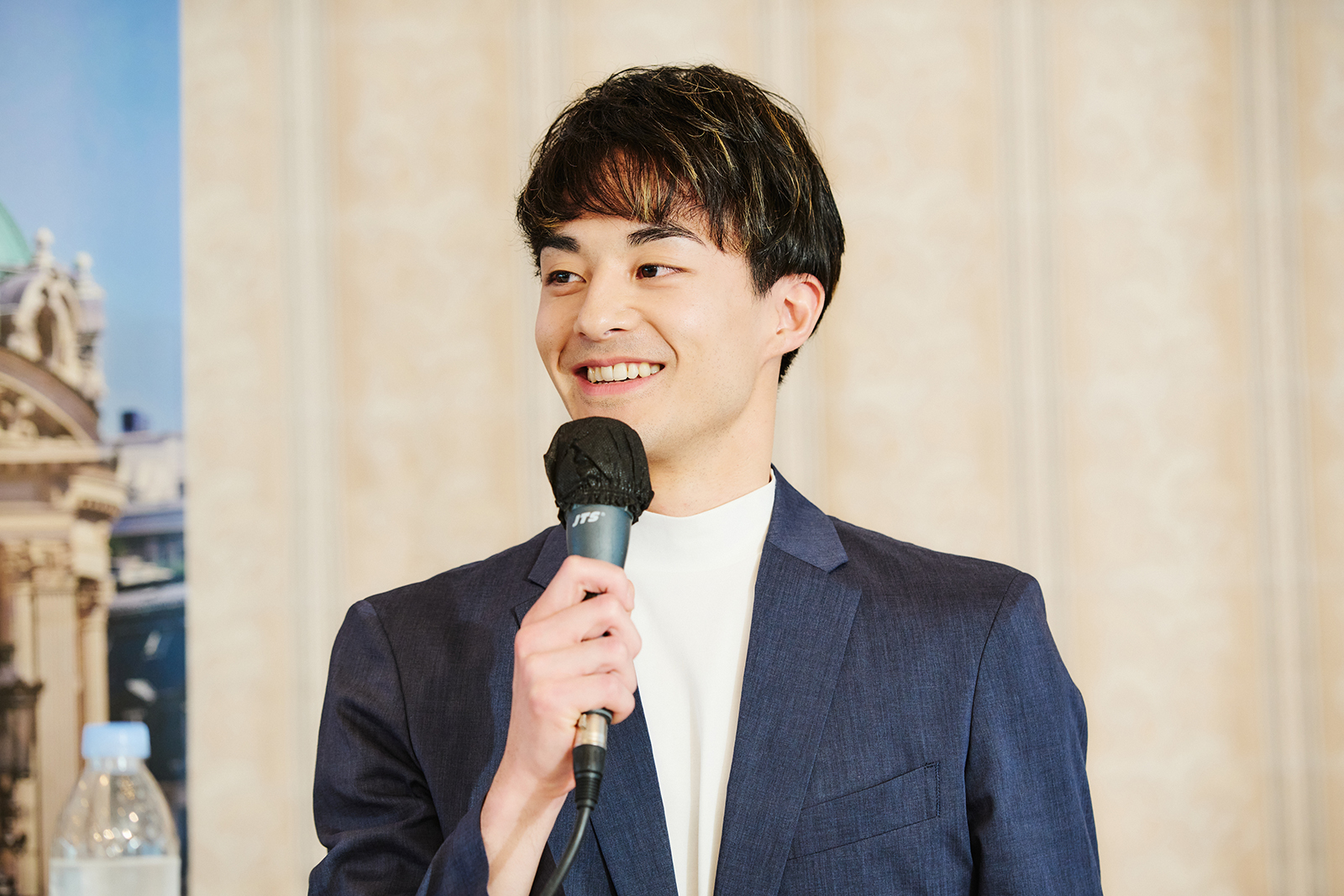 Man in a navy blazer speaks into a microphone, smiling during a presentation.