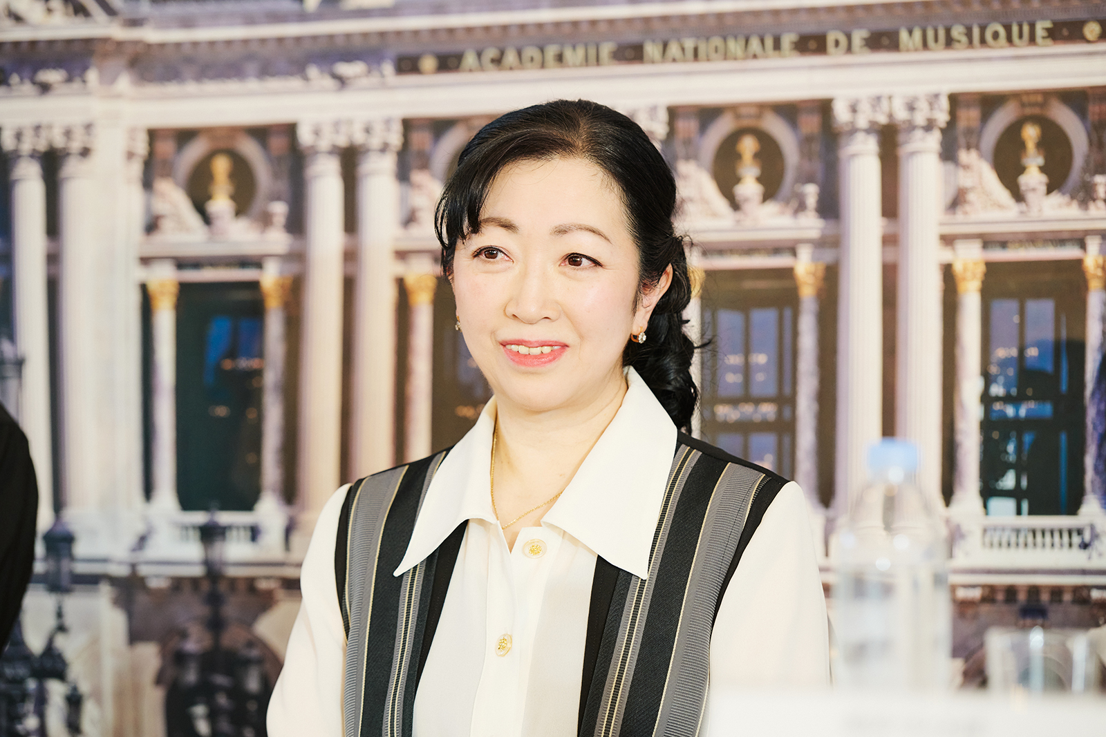 Smiling Asian woman in a white blouse with a striped vest, posed in front of a grand neoclassical building backdrop.