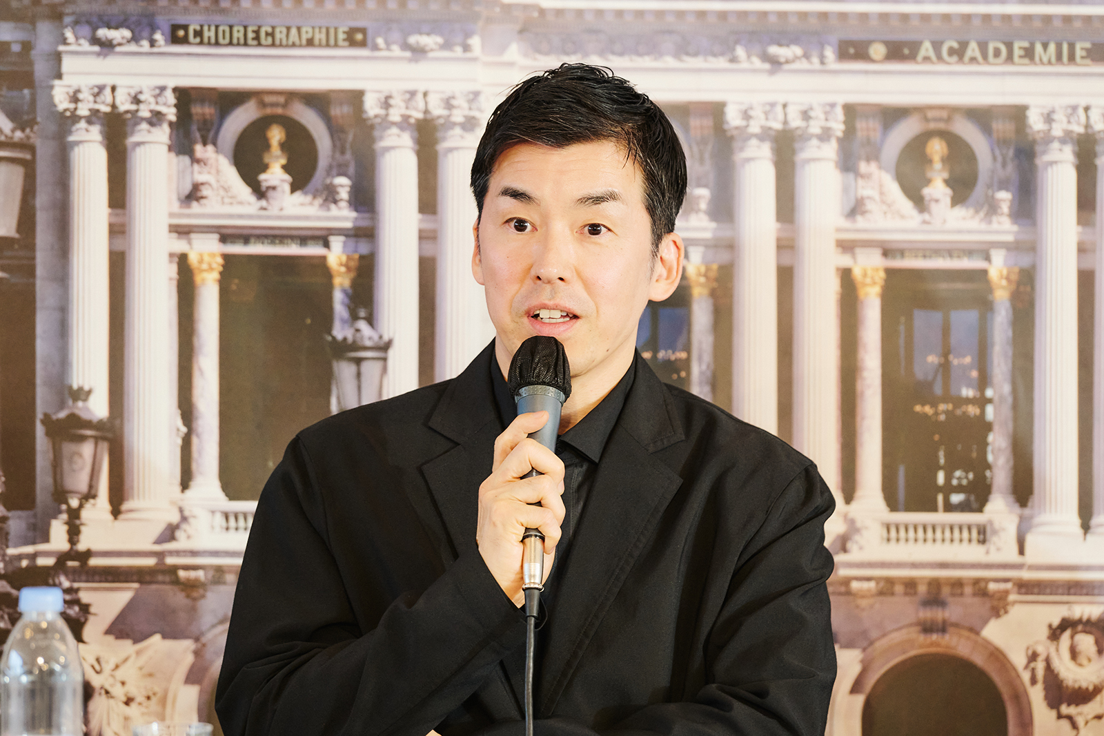 Man in a black suit speaks into a handheld microphone against a backdrop of ornate classical columns and architecture.