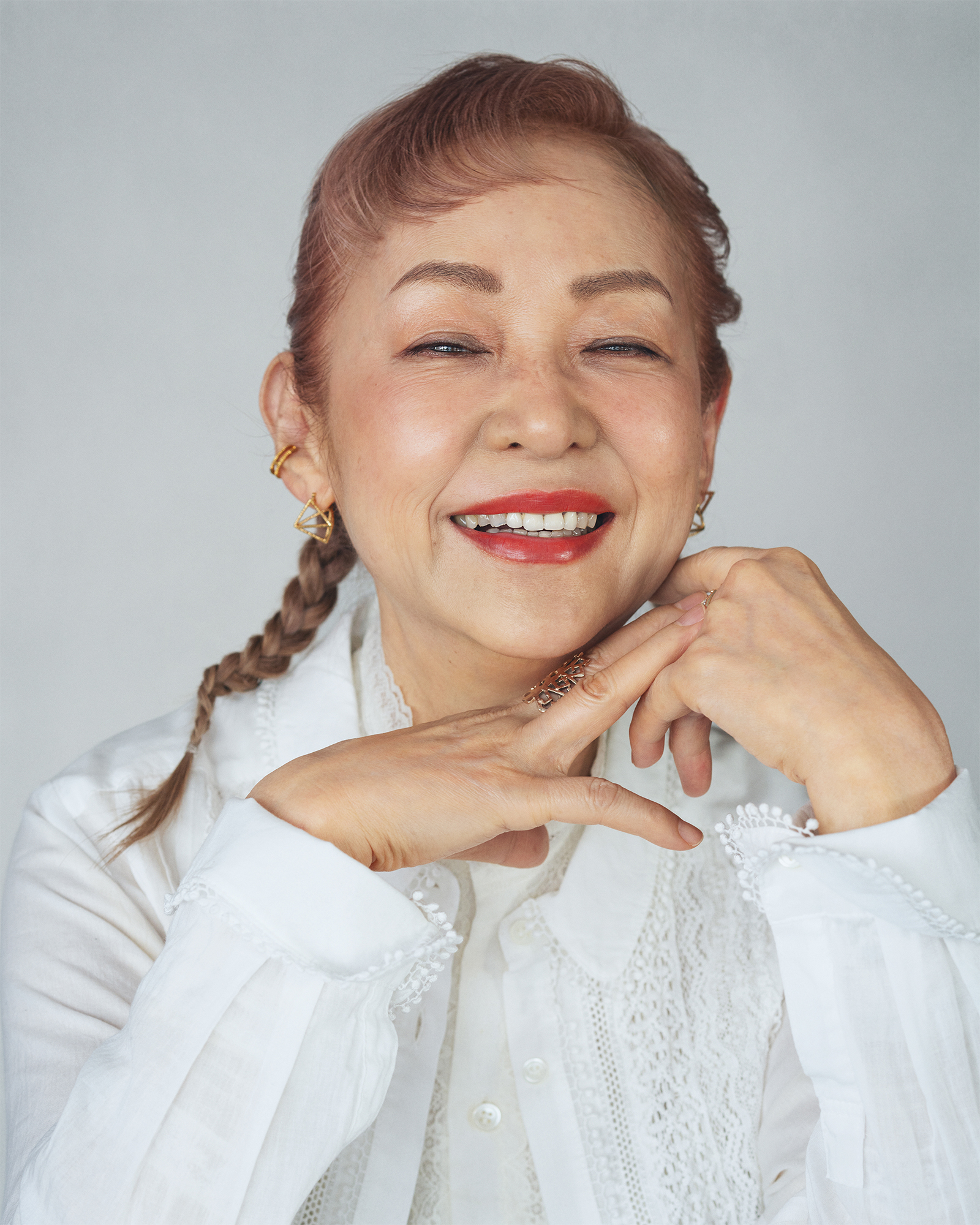 Smiling woman with braided hair wearing a white lace blouse, hands resting under her chin against a gray background.