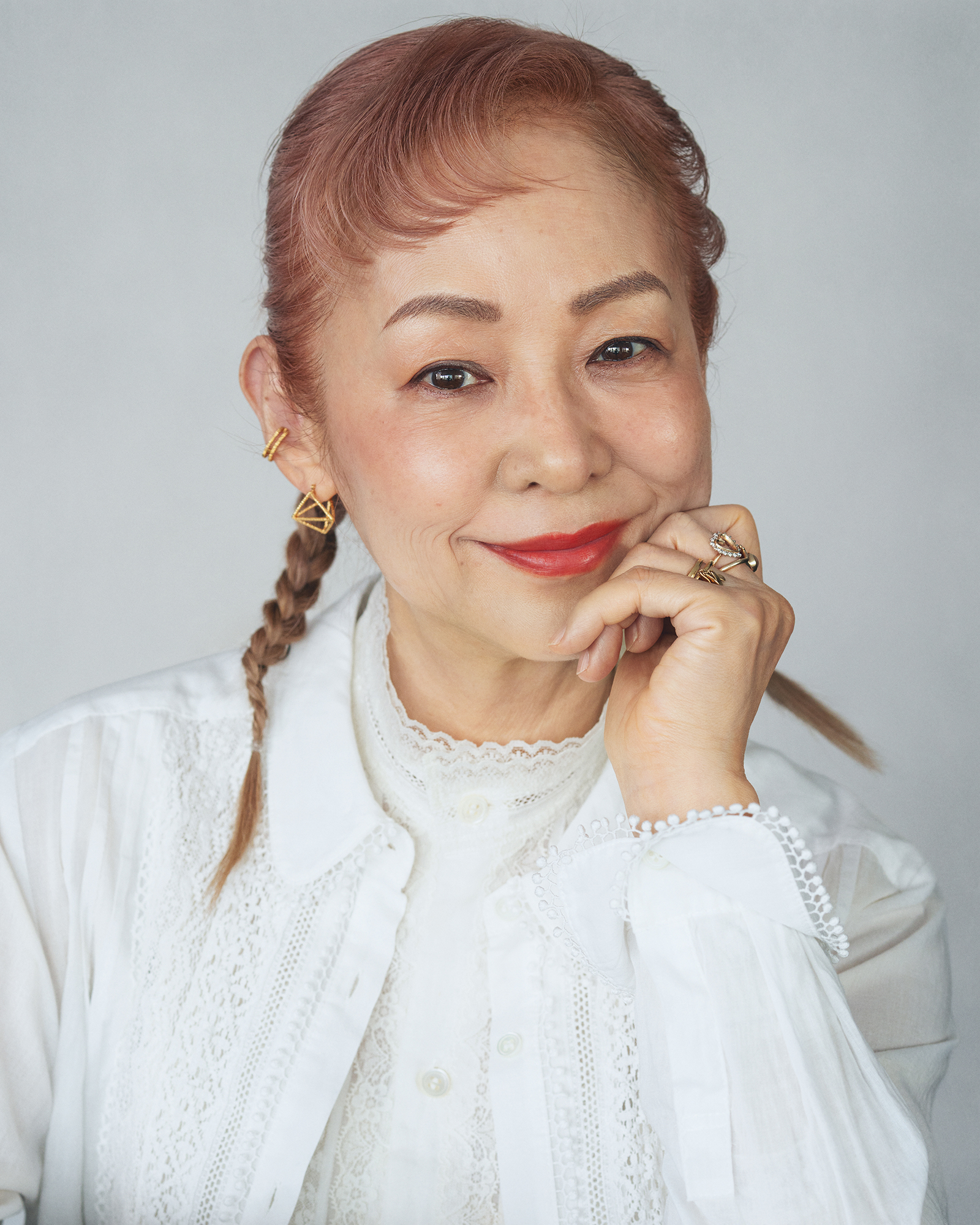 Older woman with pink hair in braids, wearing a white lace blouse, smiling softly and resting her chin on her hand.