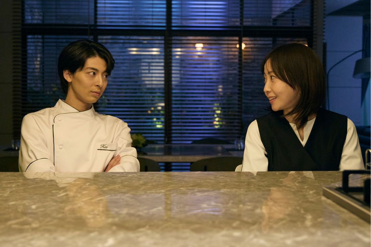Two restaurant staff sit at a marble counter, chatting: a male chef in a white coat on the left and a female server in a black vest on the right, nighttime ambiance behind them.
