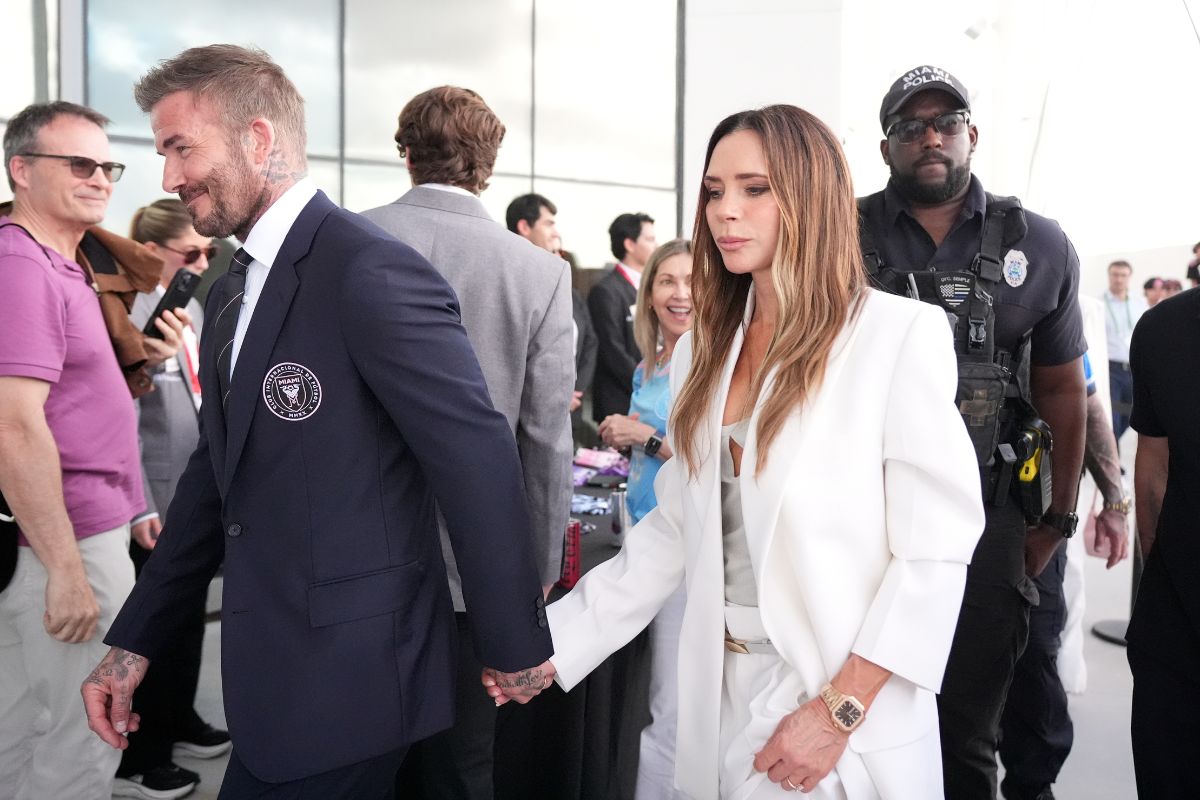 Man in a navy suit and a woman in a white blazer walk hand in hand at a crowded event, with security in the background.