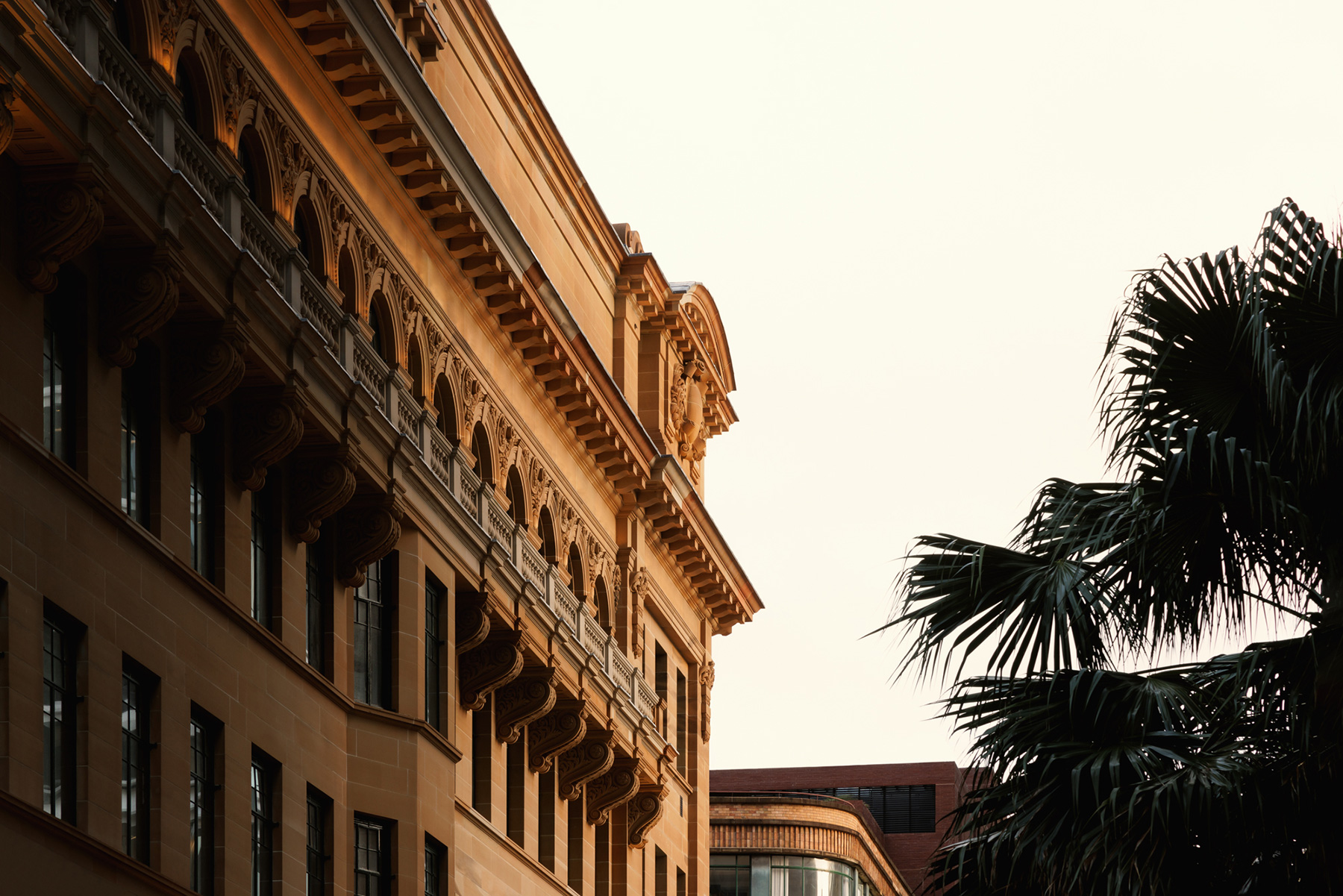 Facade of a historic sandstone building with ornate arches and decorative brackets, bathed in warm sunlight, with a palm tree on the right against a pale sky.