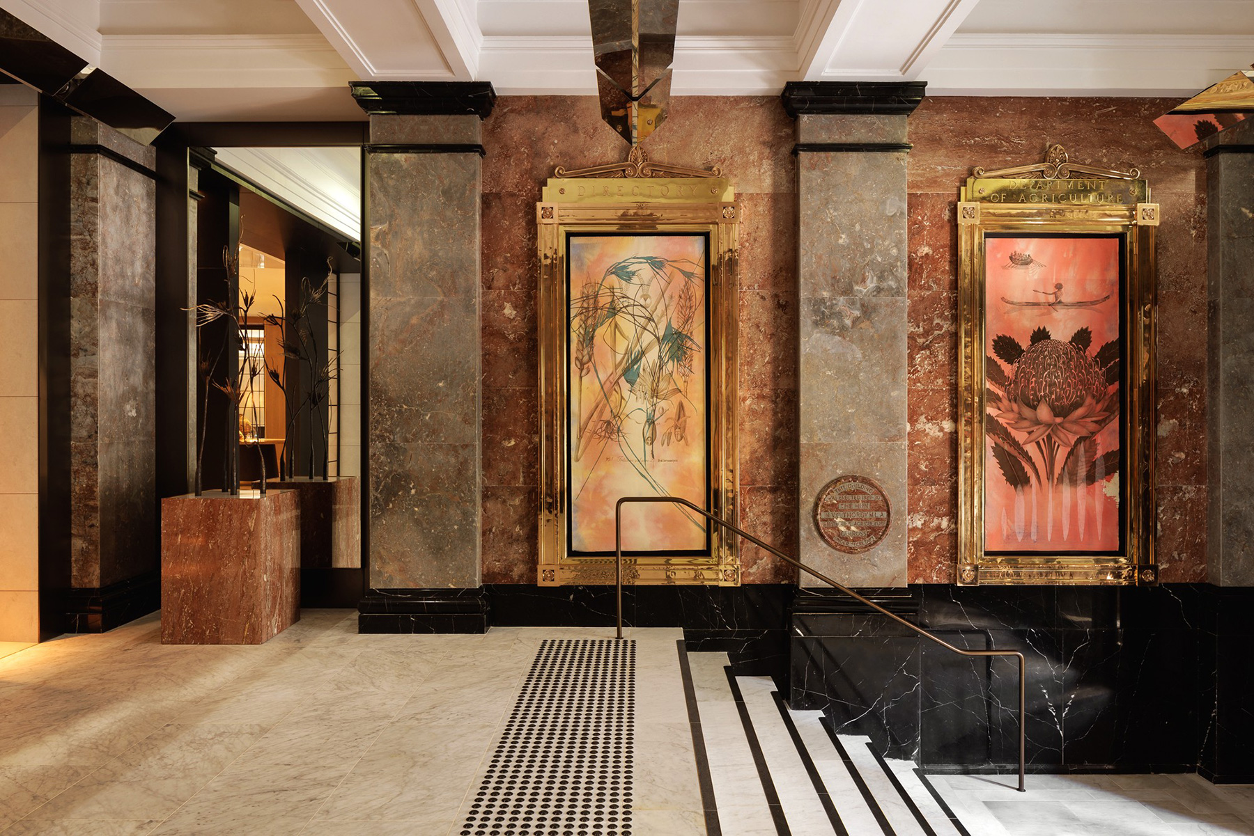 Grand marble lobby with two large, ornate gold-framed artworks flanking the center stairs and a black-and-white striped ramp leading up the steps.