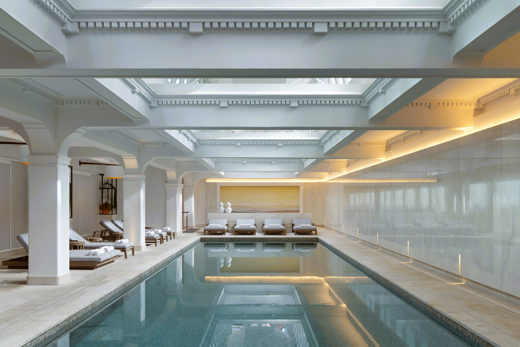 Indoor luxury spa with a long rectangular pool, white columns, and lounge chairs along the deck