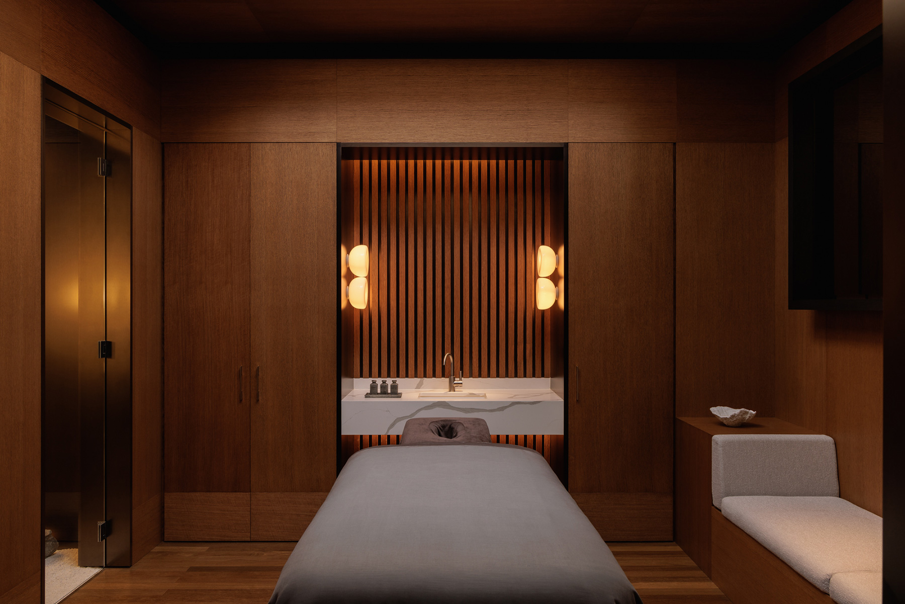 Warm, wood-paneled spa treatment room with a massage bed and a marble sink area at the head of the bed, glowing wall sconces.