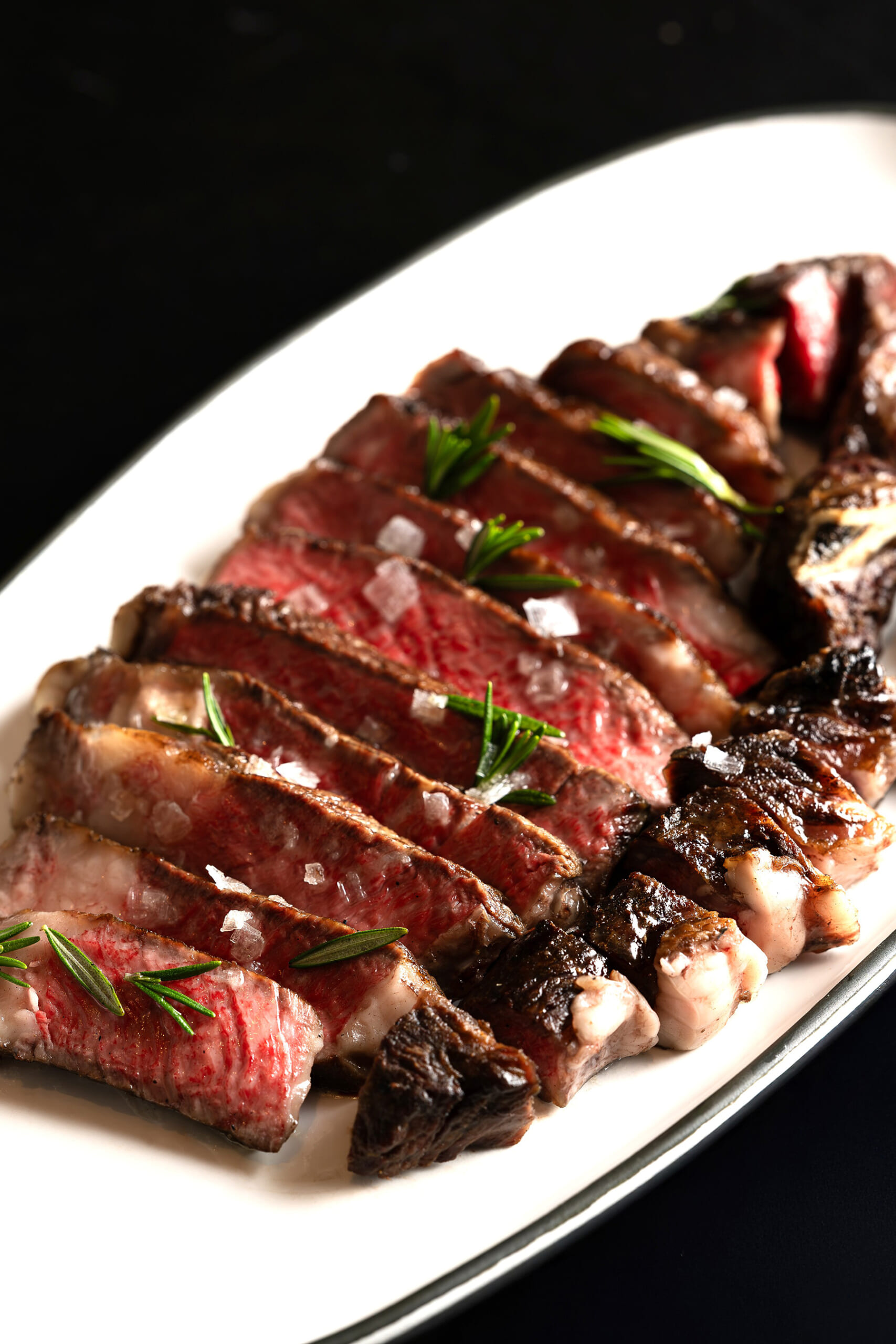 Sliced medium-rare steak arranged on a white platter, garnished with rosemary and sprinkled with coarse sea salt.