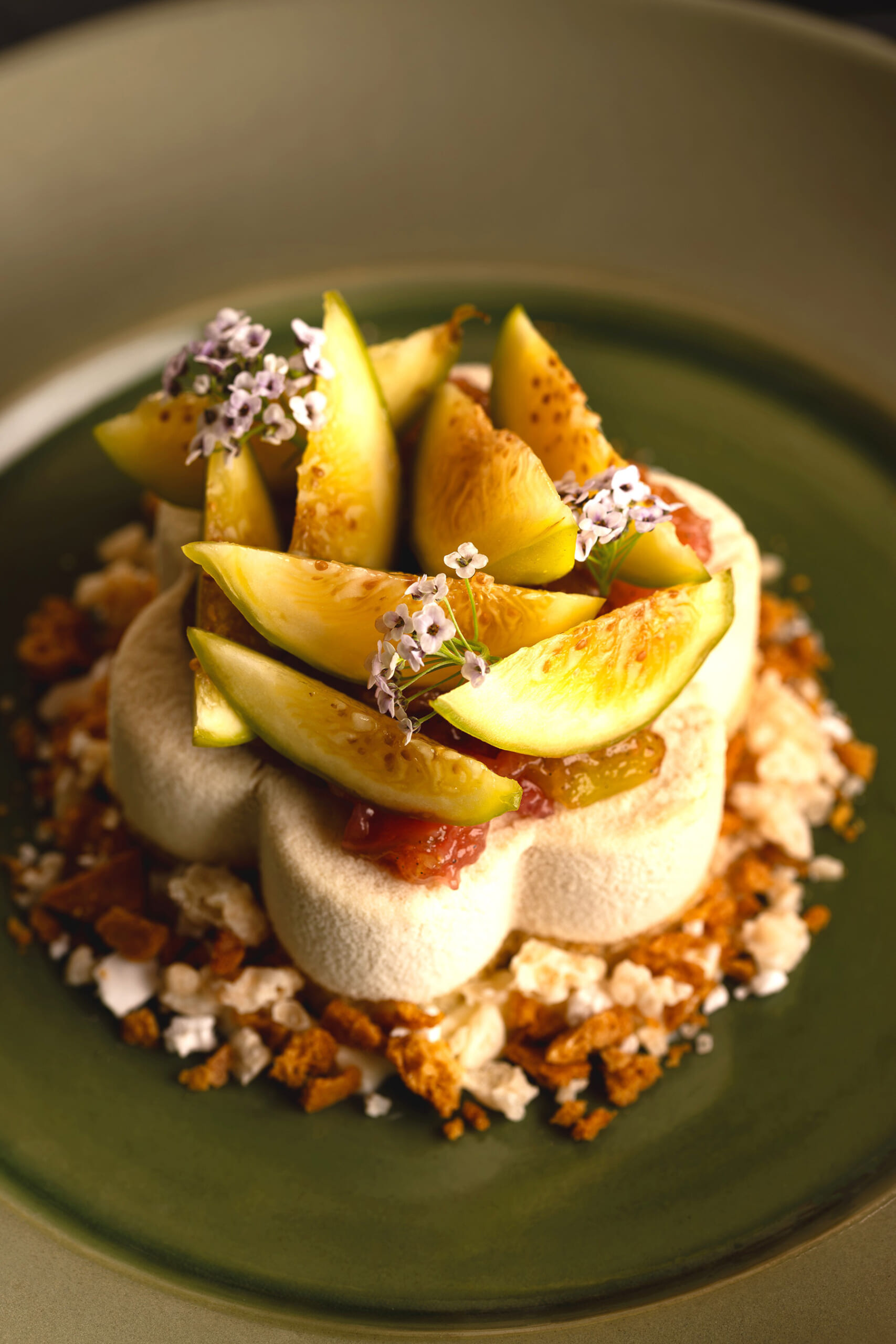 Elegant dessert: meringue ring topped with peach slices and edible flowers on a green plate, with crumbled biscuit around.