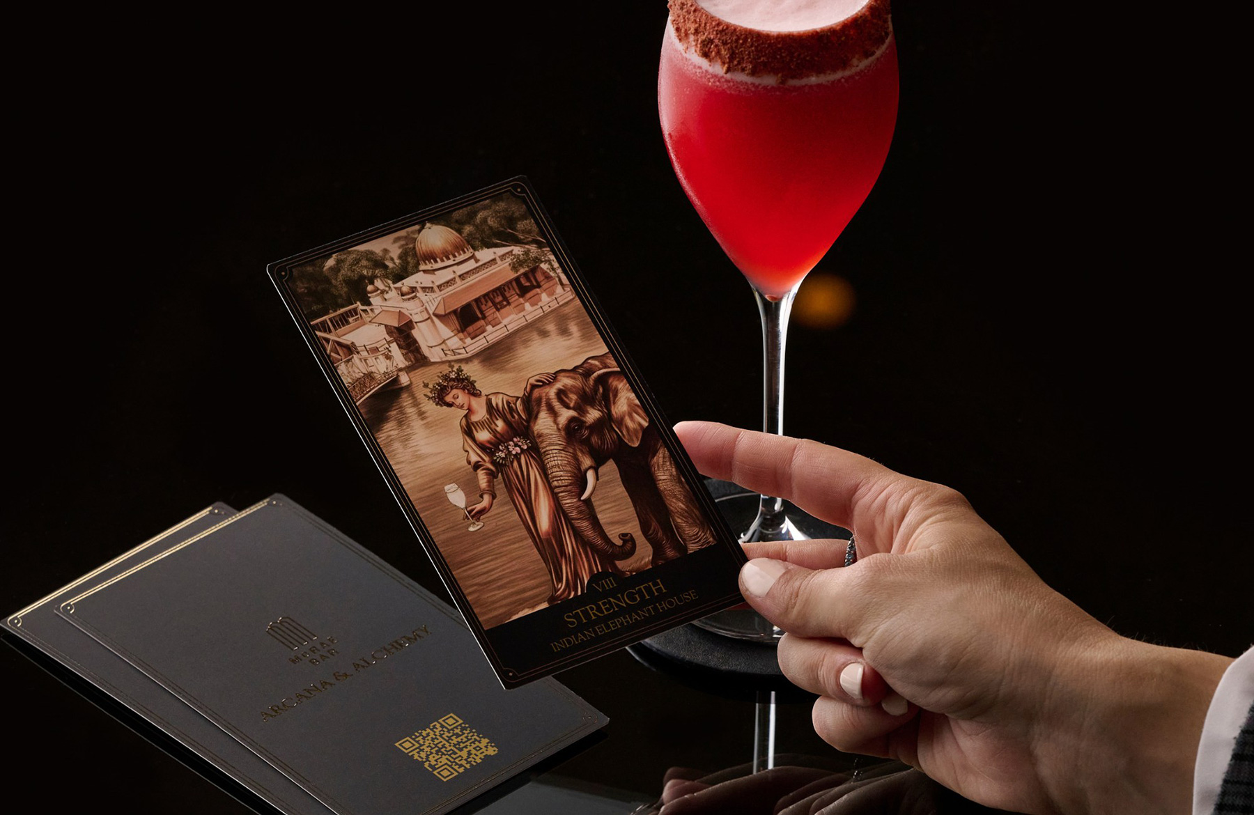 Tarot card labeled Strength, showing an elephant and a woman by water, held by a hand beside a red cocktail on a dark surface.