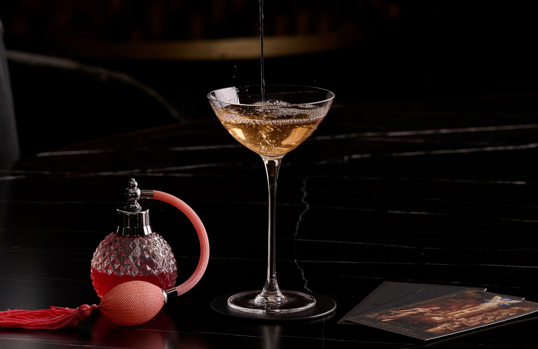 A champagne coupe is being filled with bubbly on a dark bar, beside a pink faceted perfume bottle and a few photos on the counter.