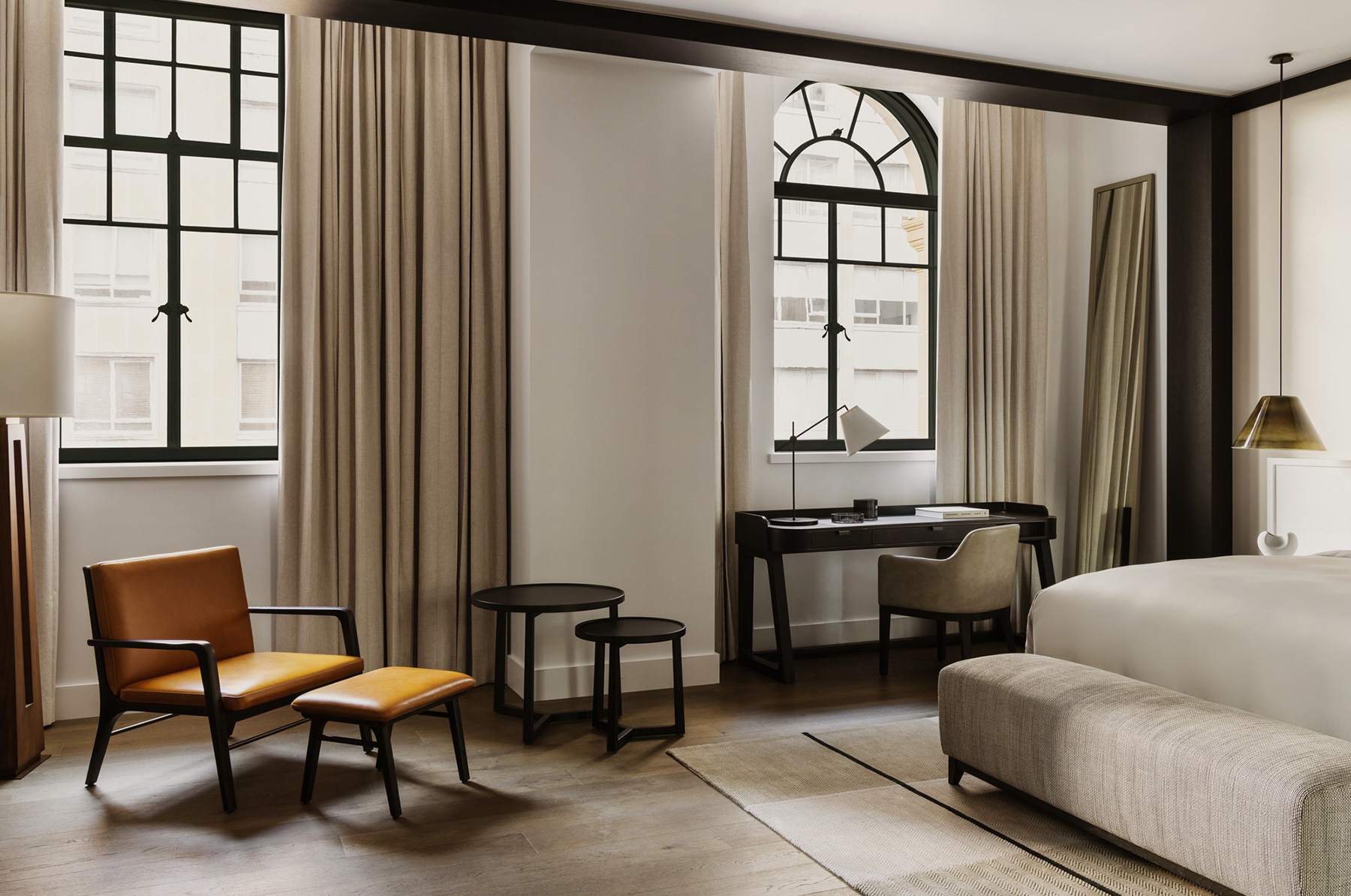 Modern hotel room living area with a tan leather chair and matching ottoman, a desk by tall windows, and a bed nearby.
