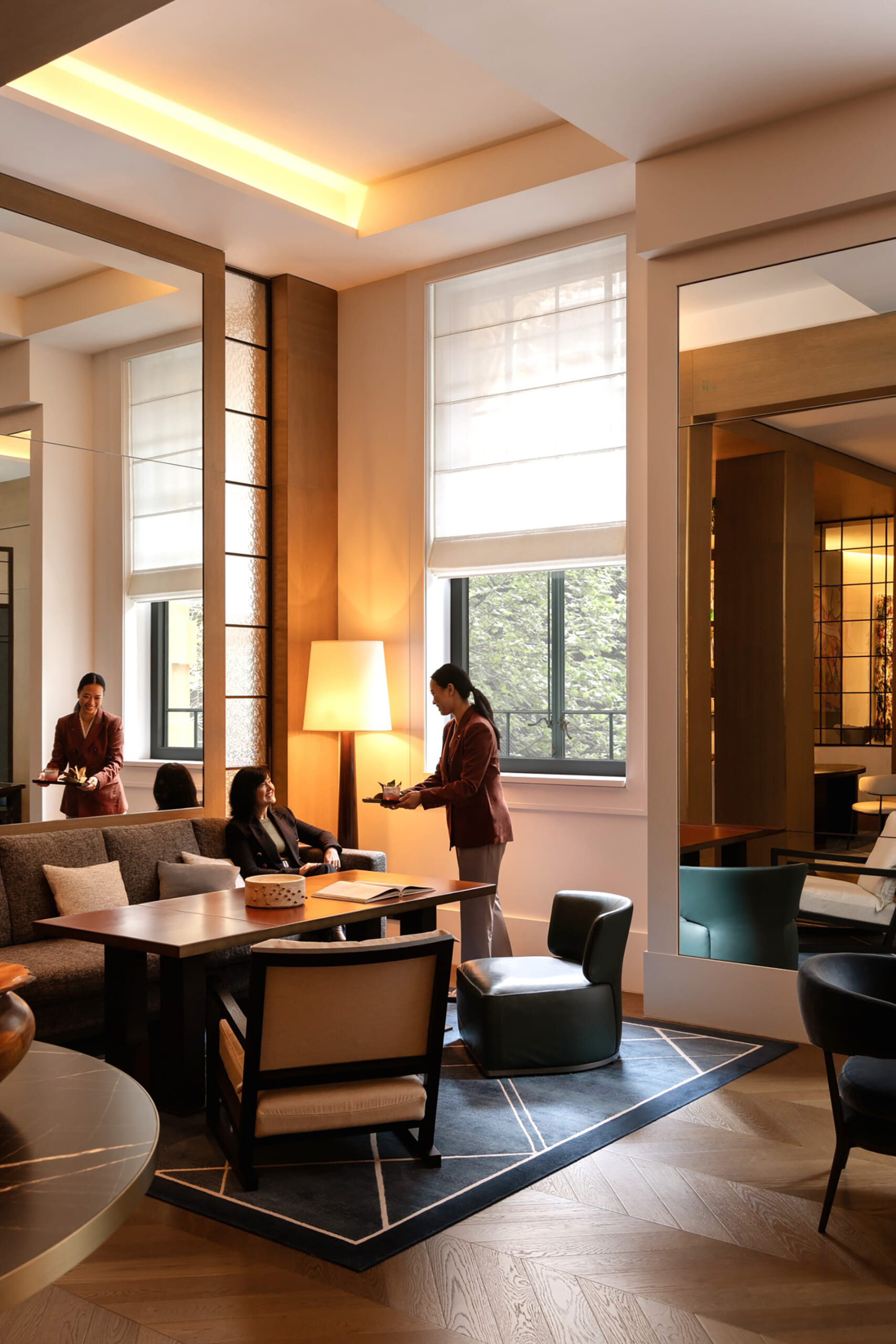 Hotel lounge with a seated guest being served by two staff near a window and sofa seating area.