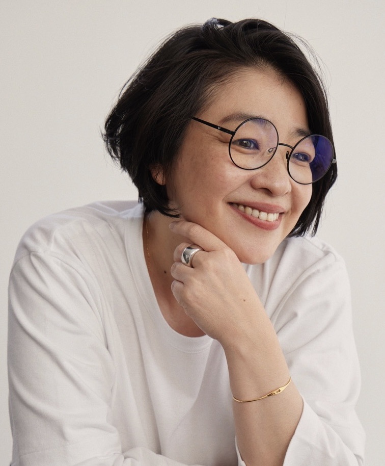 Smiling woman with short black hair and round glasses resting her chin on her hand in a white top.