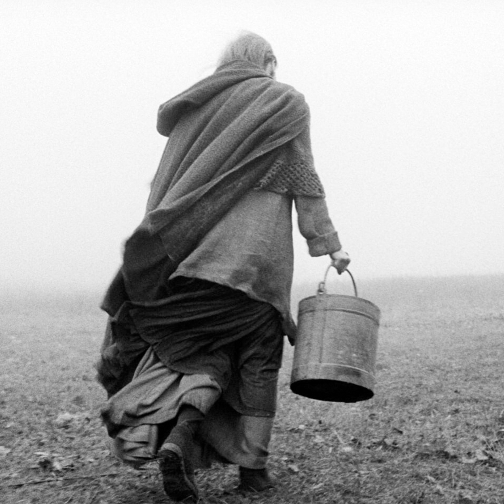 Person walking away across a barren field, wrapped in a heavy cloak and carrying a metal bucket by its handle.