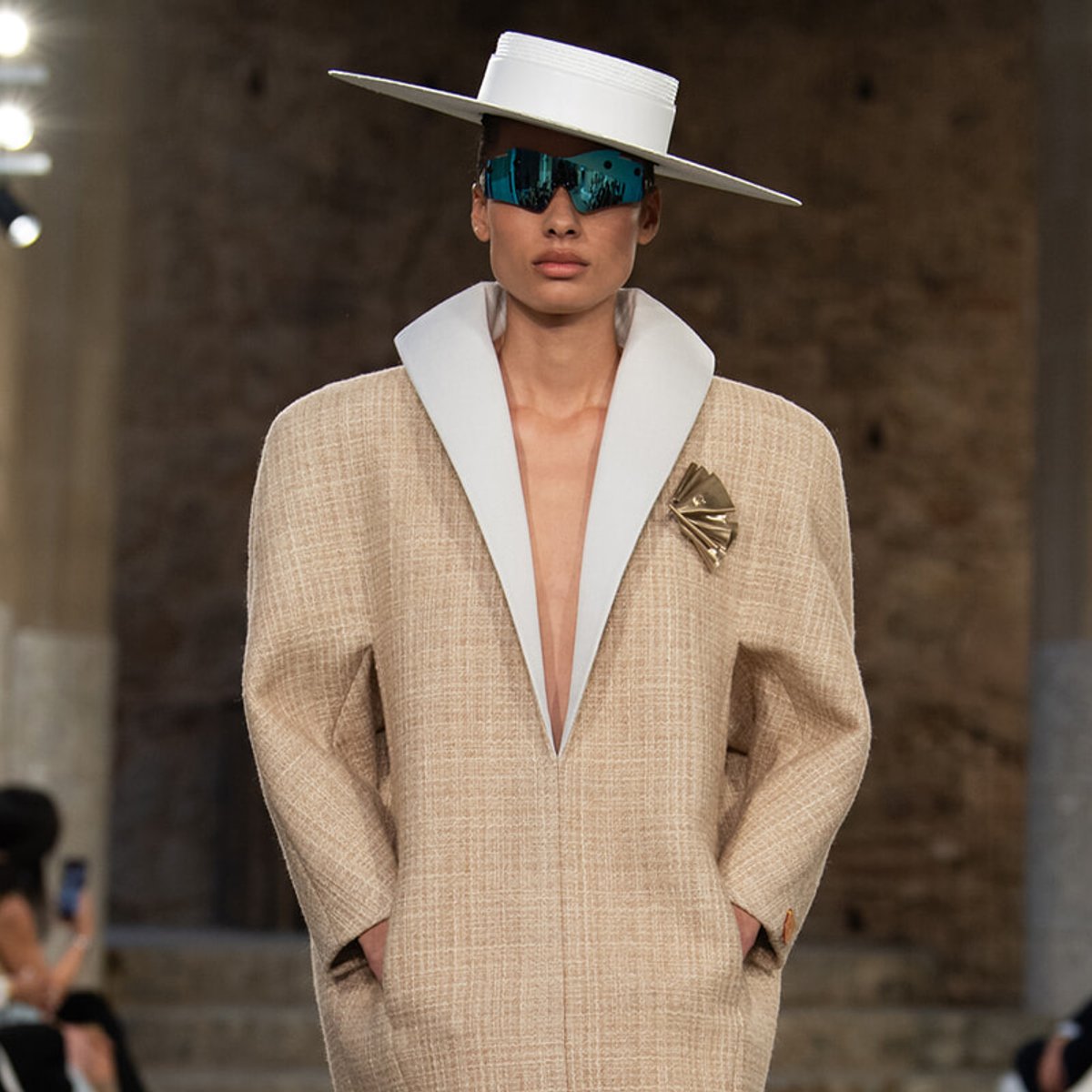 Model on a runway wearing an oversized beige coat with white lapels, a white wide-brim hat, blue mirrored sunglasses, and a gold brooch ornamental pin.