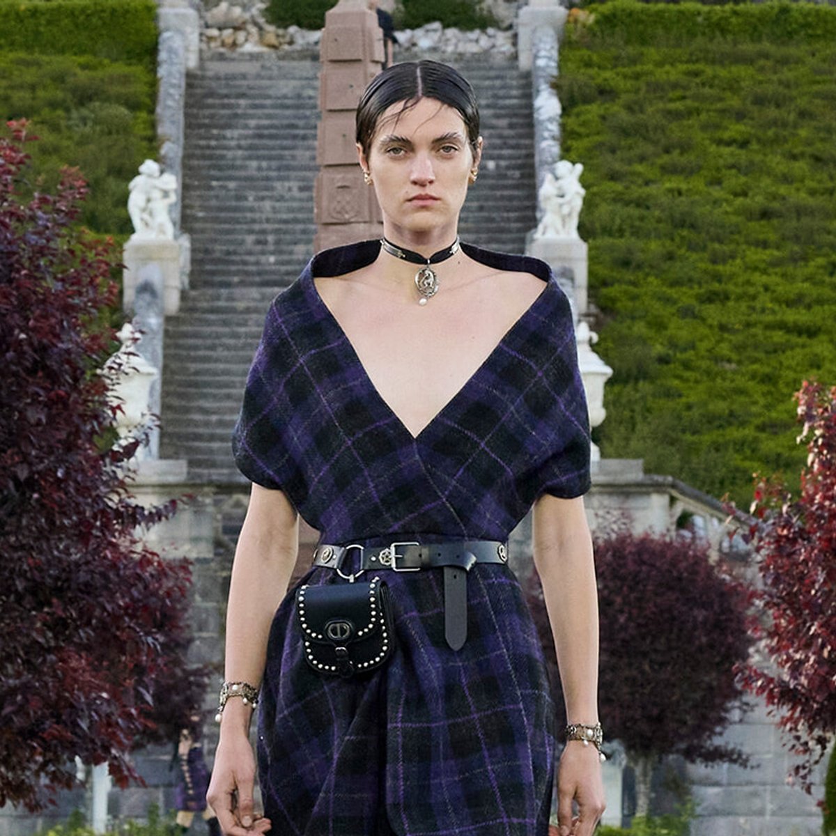 Woman in a purple plaid wrap dress with a black belt and studded pouch, standing before a stone staircase outdoors, garden setting.