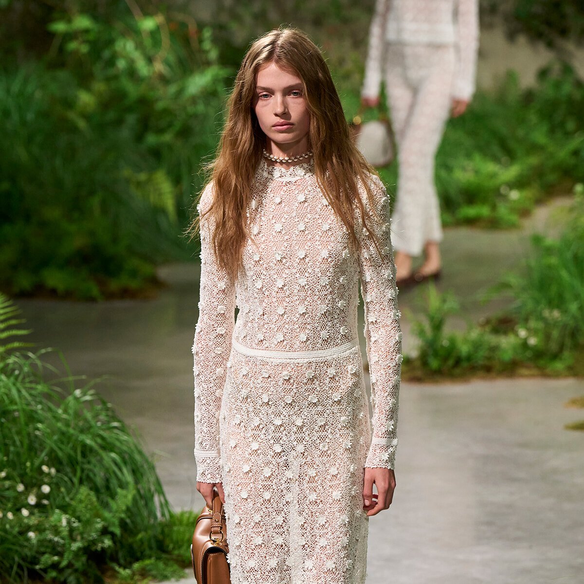 Model walking a runway in a long white lace dress with floral dots, holding a brown handbag in a green garden setting