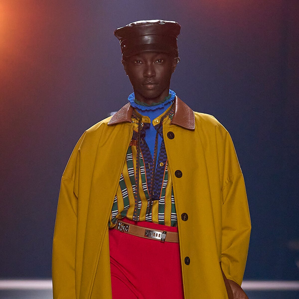 Model walking the runway in a bright yellow coat over a multicolored striped shirt, blue turtleneck, red trousers, and a black cap.