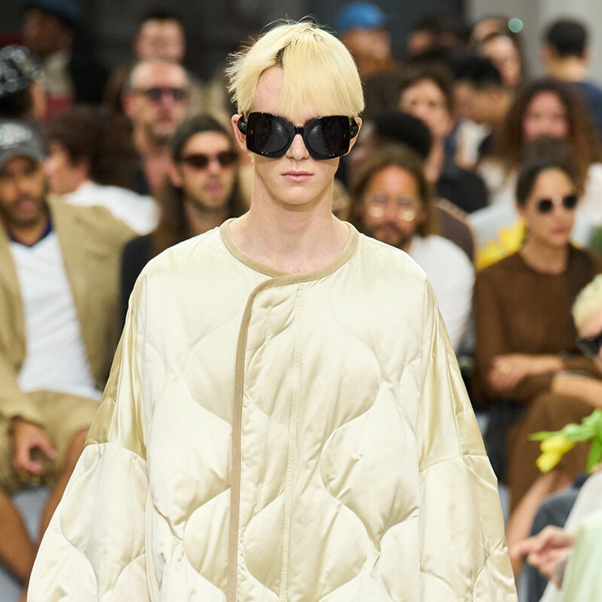 Model on a runway wearing a cream quilted oversized coat and large black sunglasses, with blonde hair and a blurred crowd behind.