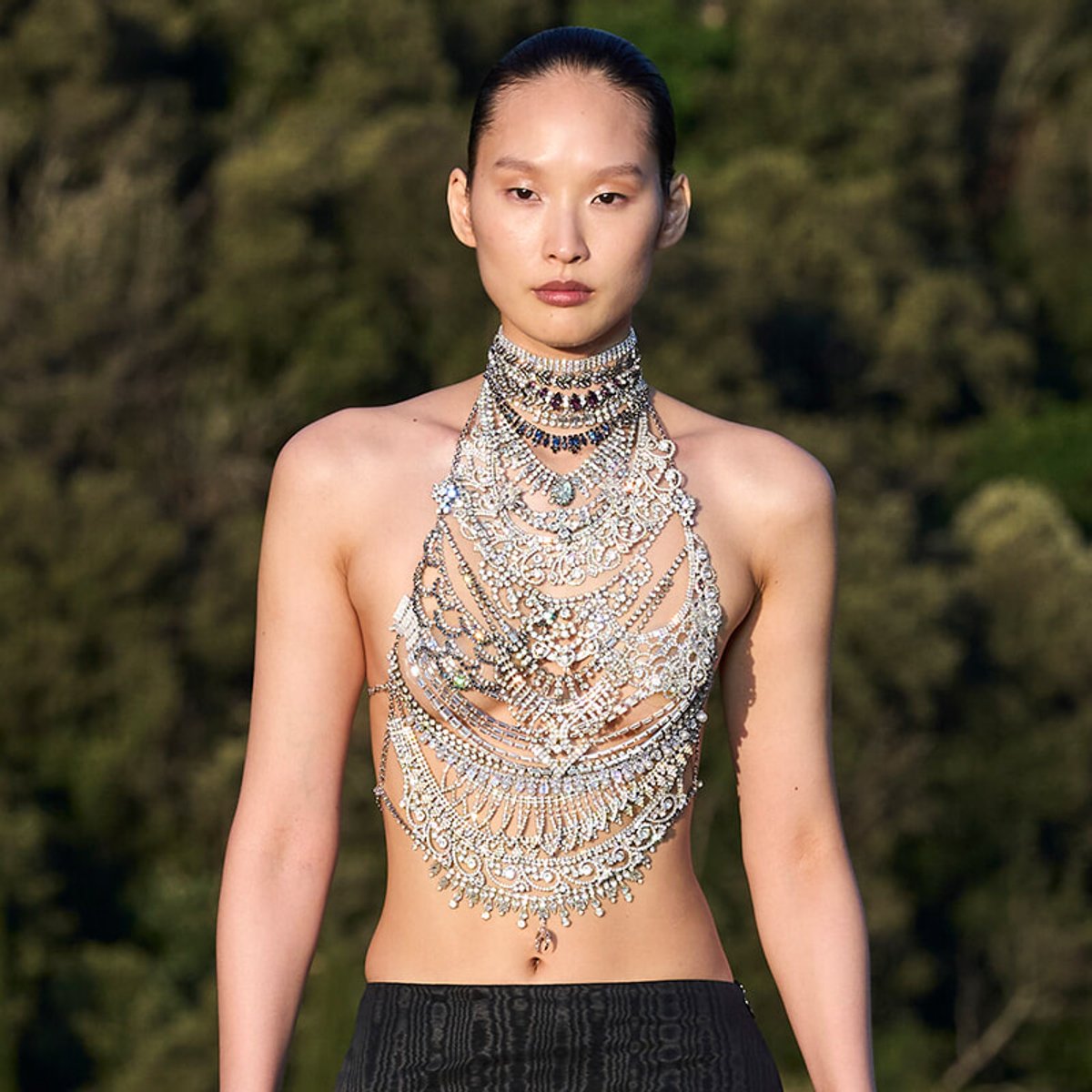 Model wearing an ornate multi-layered crystal necklace draped across the chest outdoors, bare shoulders visible.