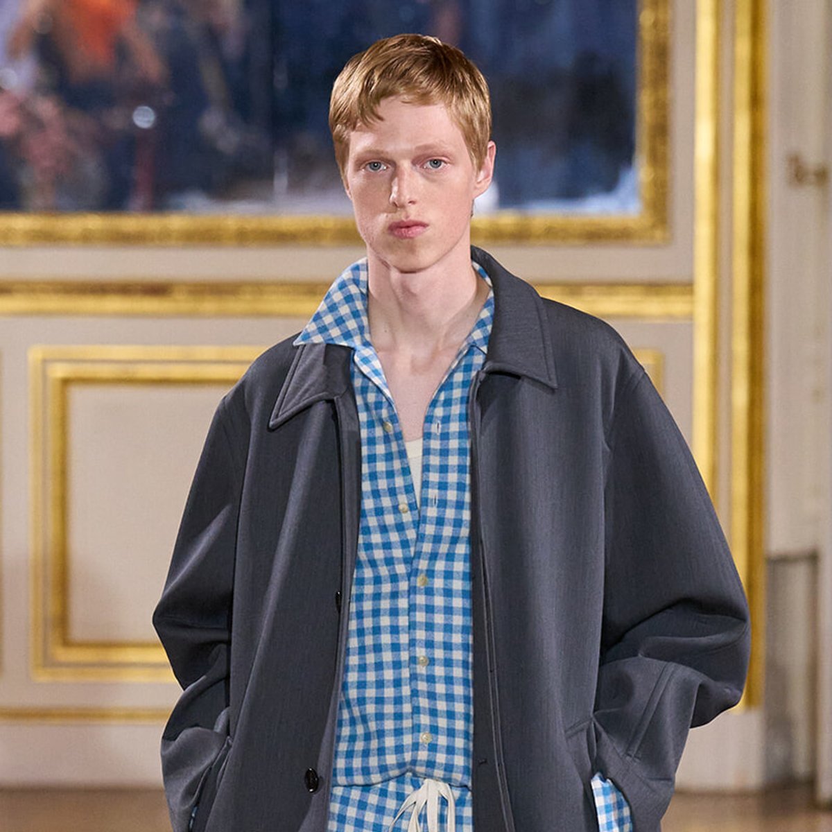 Young man in a blue checkered shirt under a dark oversized coat stands in a room with gold-framed walls or paintings behind him.