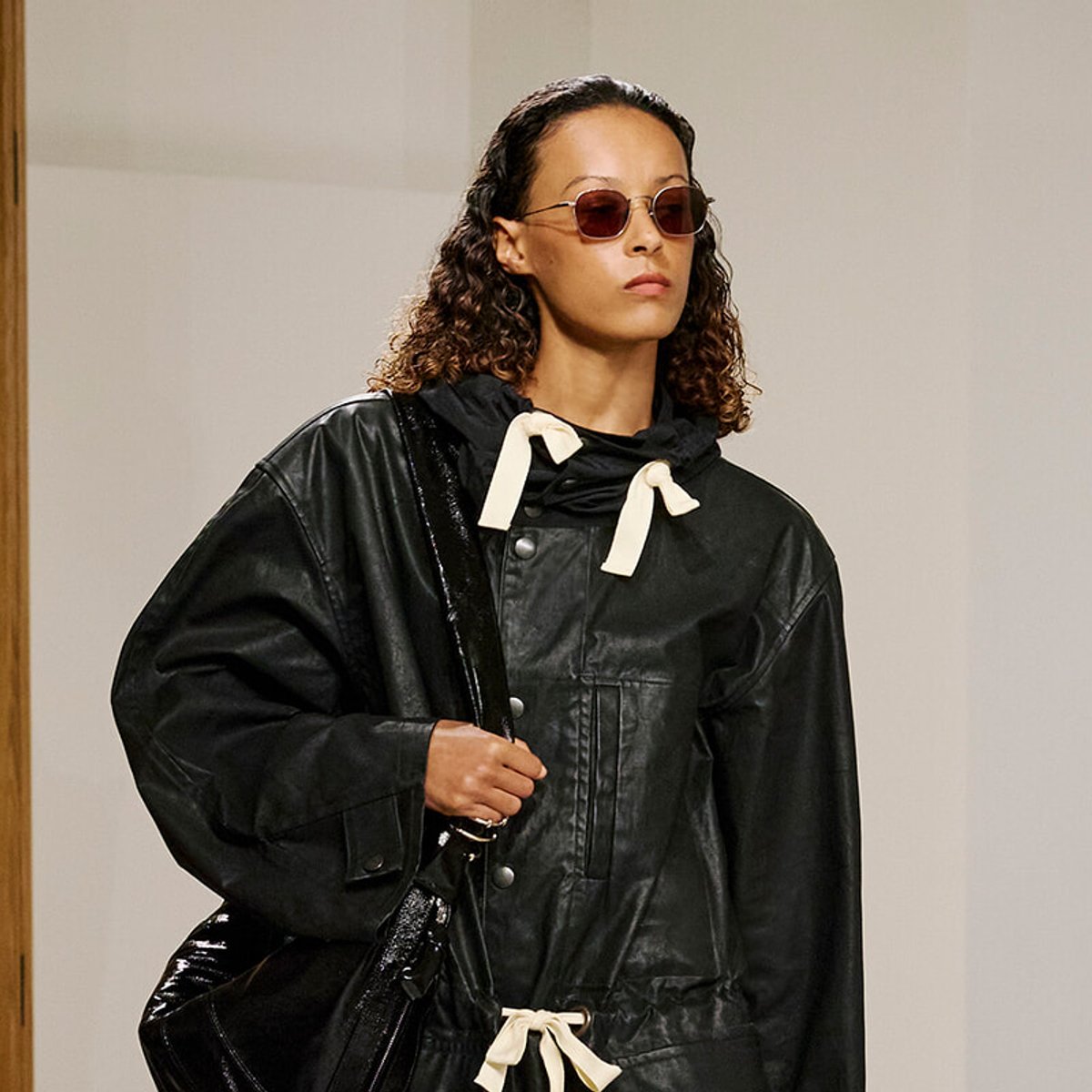 Model wearing a black glossy rain jacket with cream drawstrings, sunglasses, and a black bag over the shoulder.