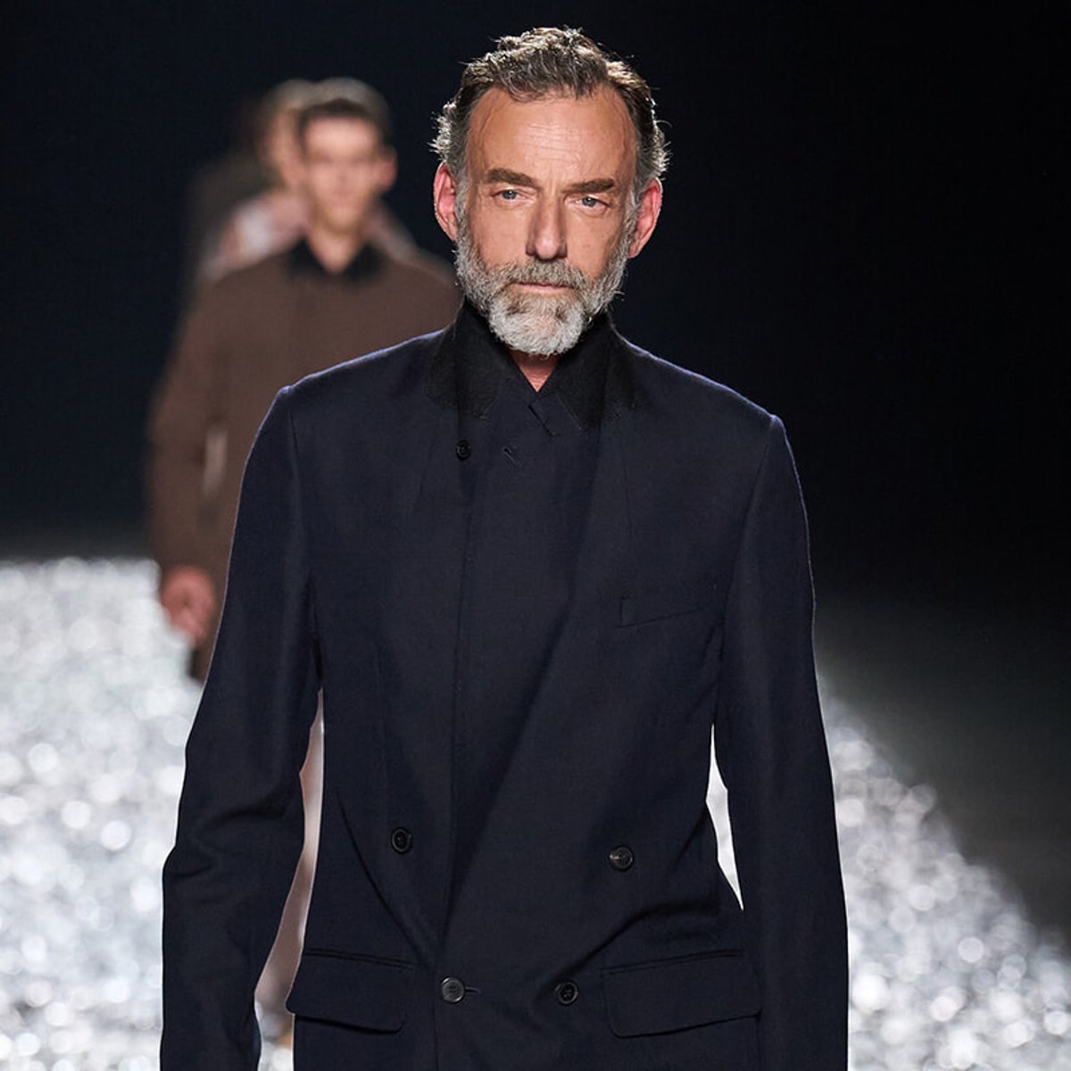 Male model with gray beard walking a runway in a dark navy double-breasted blazer; glittering runway lights in the background.
