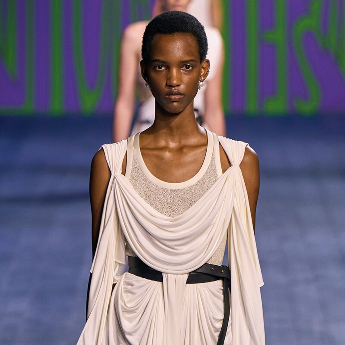 Model walking a runway in a cream draped gown with a wide belt and sheer panel at the neckline.