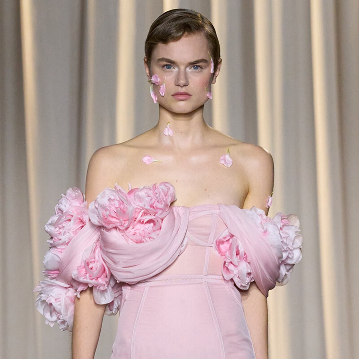 Runway model in a pale pink gown with large ruffled floral accents, petals scattered on shoulders and face.