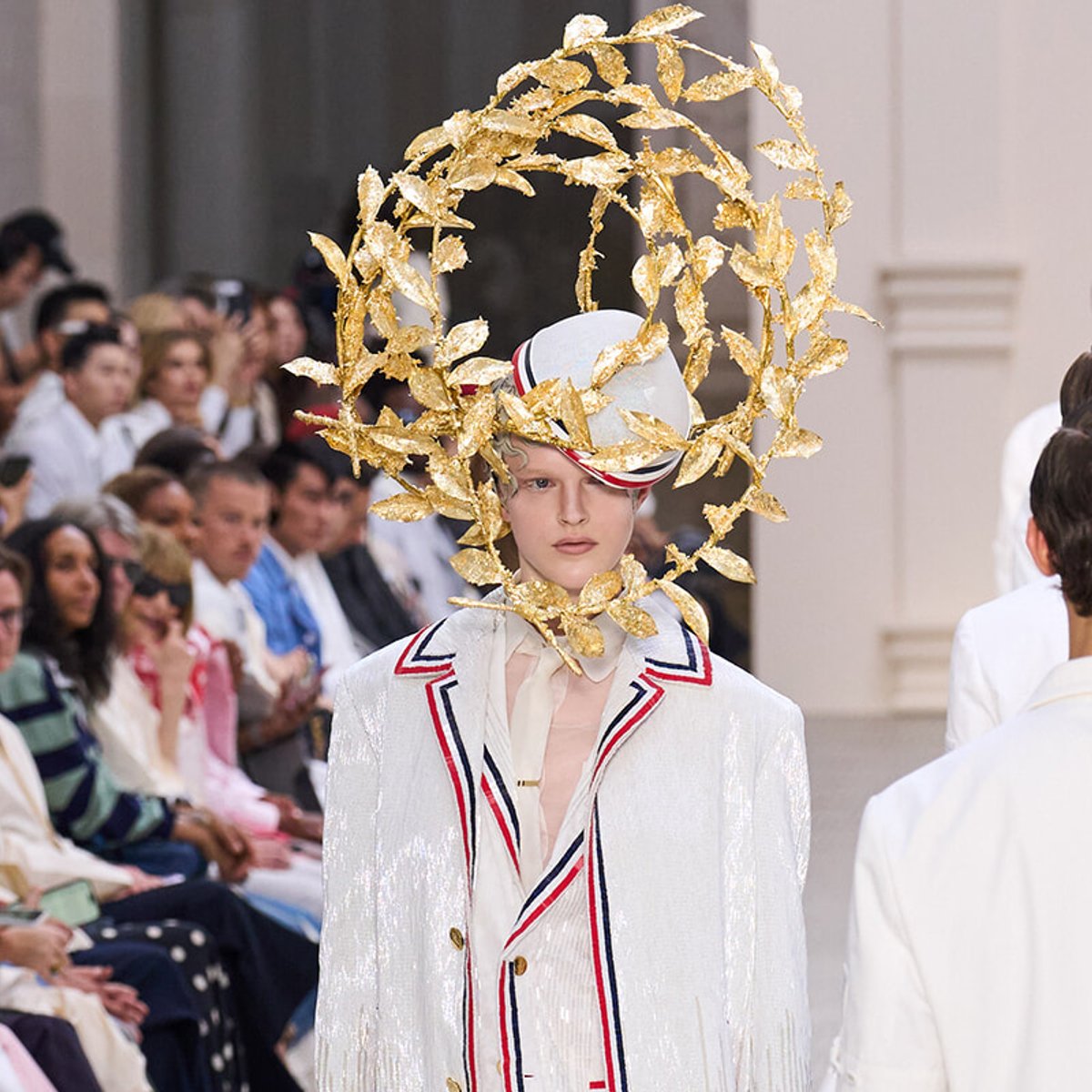 Model on a runway wearing a white suit with red/blue piping and a large gilded leaf halo headpiece framing her face.