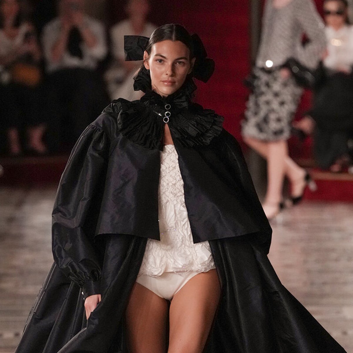 Model walking the runway in a voluminous black cape with a ruffled high collar, paired with a white lace top and white bottoms, against a fashion show backdrop.