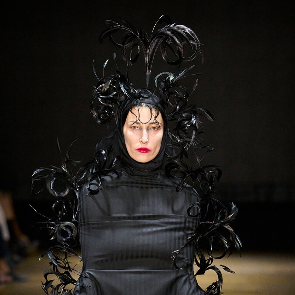 Model on a runway wearing a glossy black avant-garde dress with a tall feathered headpiece.