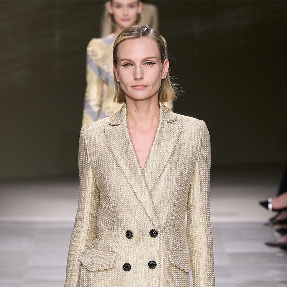 Female fashion model walking a runway in a textured cream blazer with black buttons, neutral makeup, and short blonde hair.