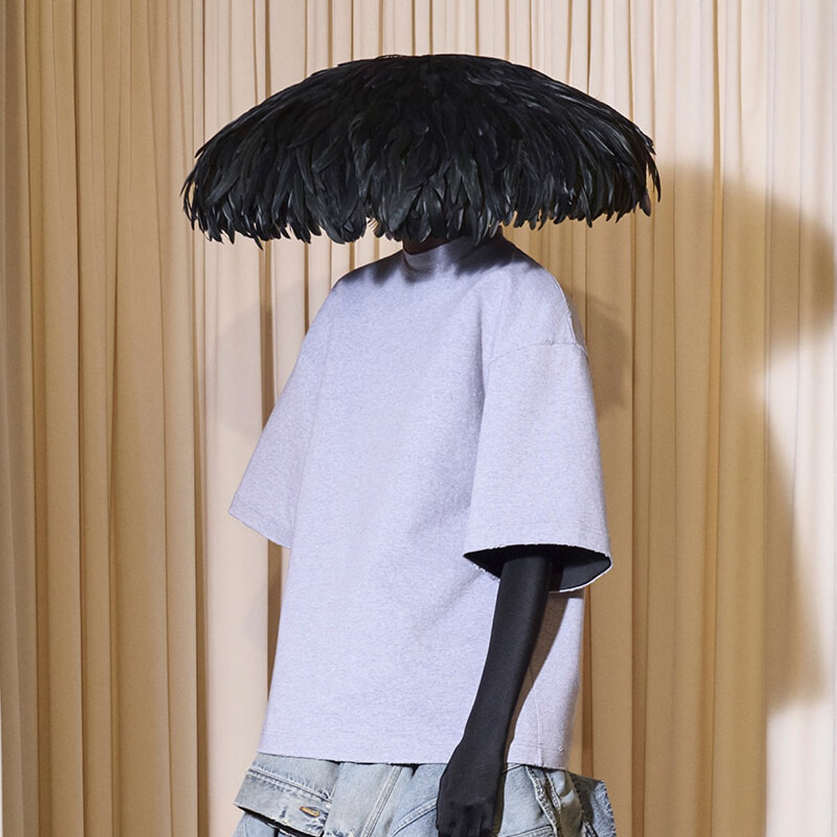 Mannequin wearing a lavender short-sleeve top and light denim shorts, with a large black feathered hat obscuring the face, against a beige curtain backdrop.