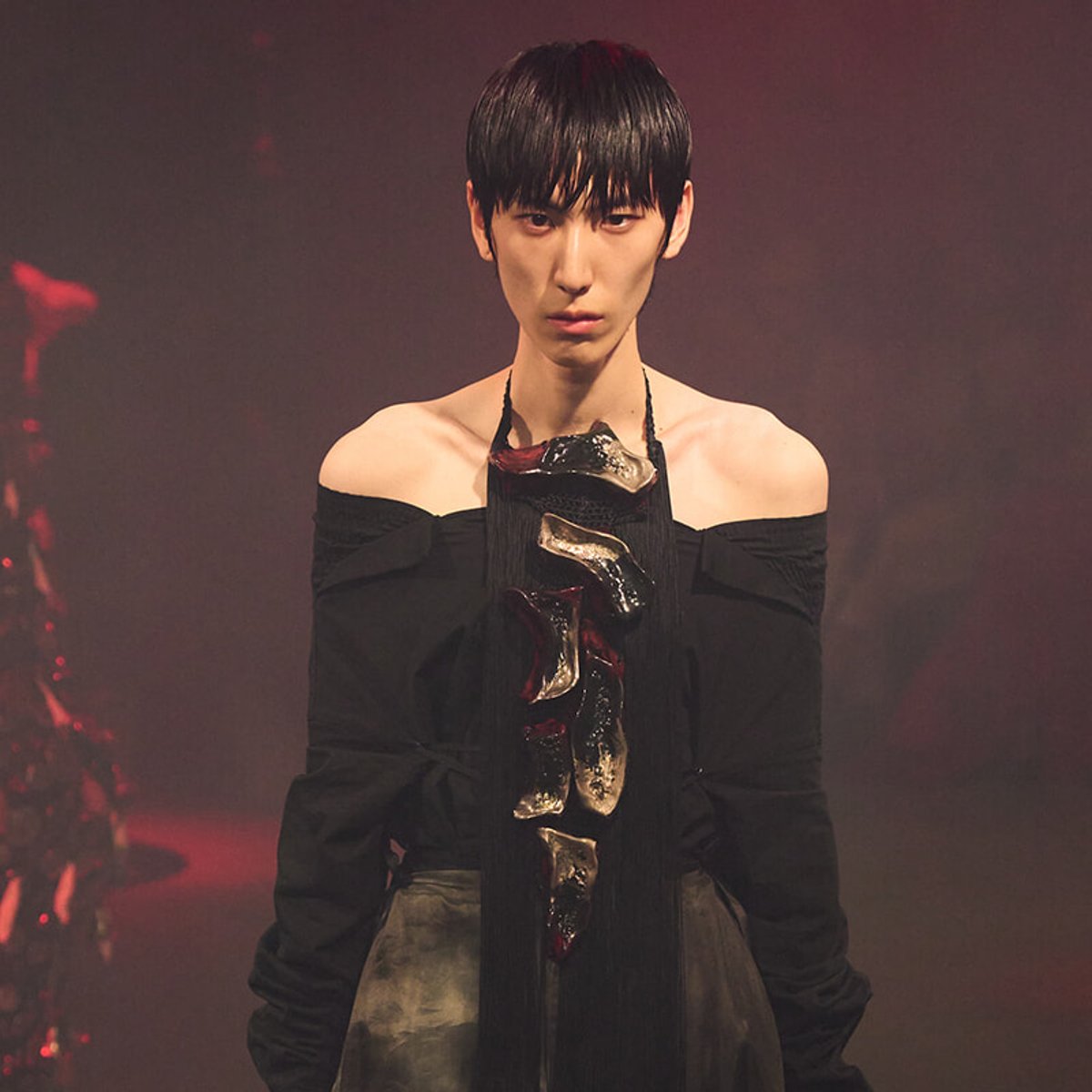 Fashion model on a dim catwalk wearing an off-the-shoulder black top and a long black scarf with metallic accents, looking forward.