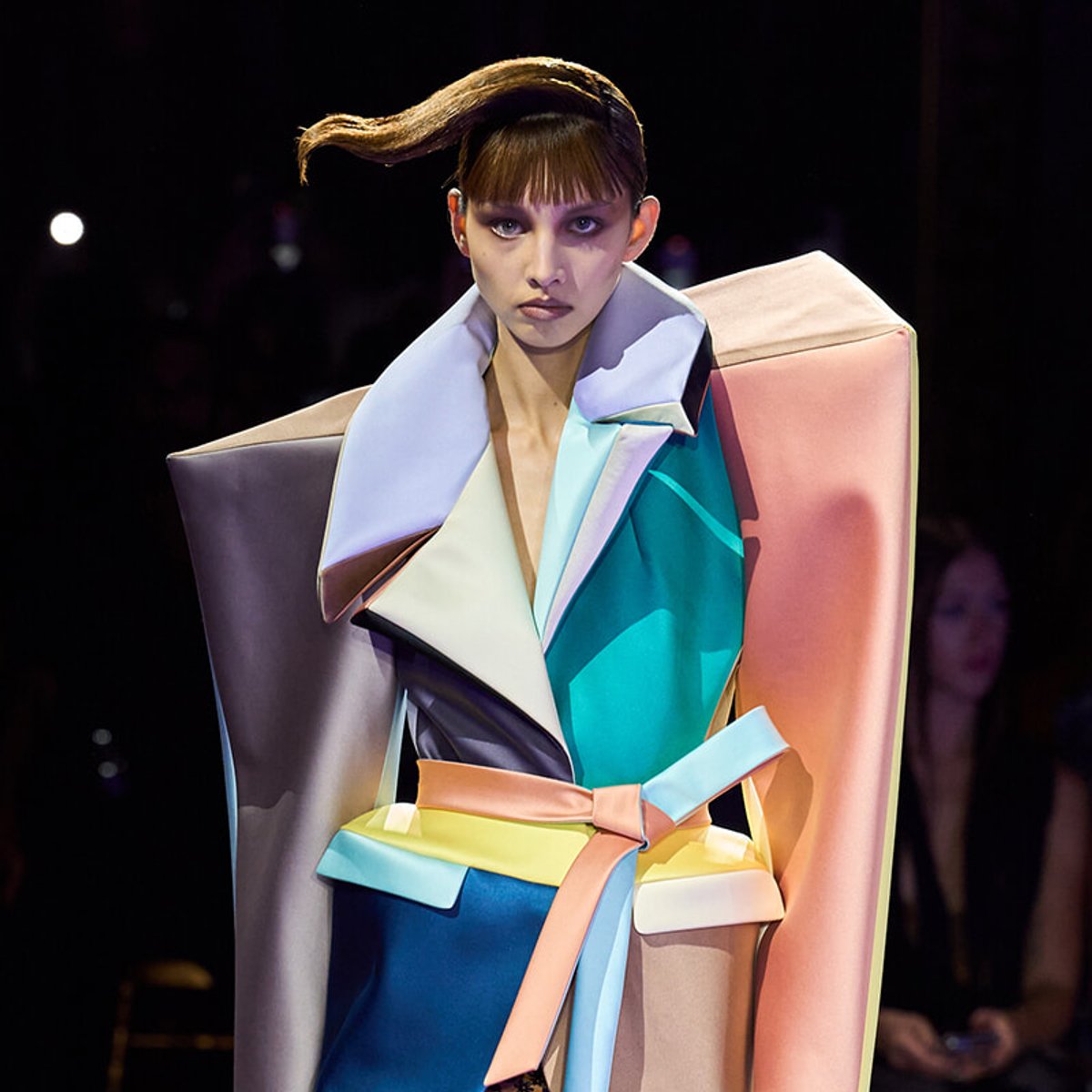 Model on a runway wearing a bold, color-blocked oversized coat with large geometric panels and a pastel belt.